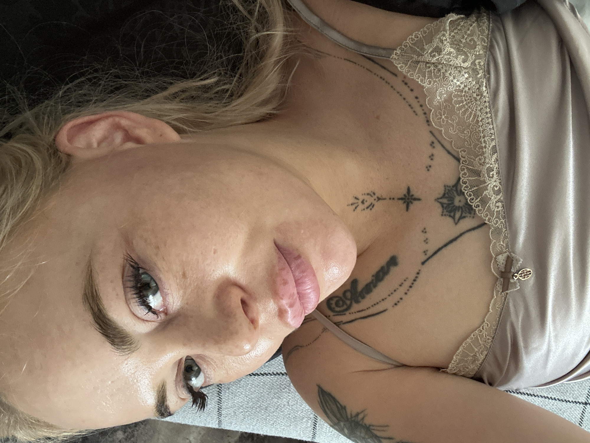 A close-up portrait shows an individual reclining, gazing towards the upper left quadrant of the frame. The person has light-toned skin with visible texture and pigmentation, full light-colored lips with a glossy finish, and light-colored eyes enhanced with prominent dark eyelashes and defined eyebrows. Blonde hair is visible around the right side of the head. The individual is wearing a light, possibly champagne or beige-colored, satin camisole featuring lace trim along the neckline. A small dark bow with a metallic, possibly gold-toned, oval charm is affixed to the left strap. Multiple tattoos are present on the upper body: a large design on the left shoulder/upper arm, script text on the right chest, and a collection of symbolic designs including a small stylized plant, a starburst, a floral pattern, and dotted lines across the chest and collarbone area. The person is positioned against a light grey textile surface with a subtle checkered pattern on the left, while the background to the right and top is dark and indistinct. The scene is located in Unknown, Unknown.