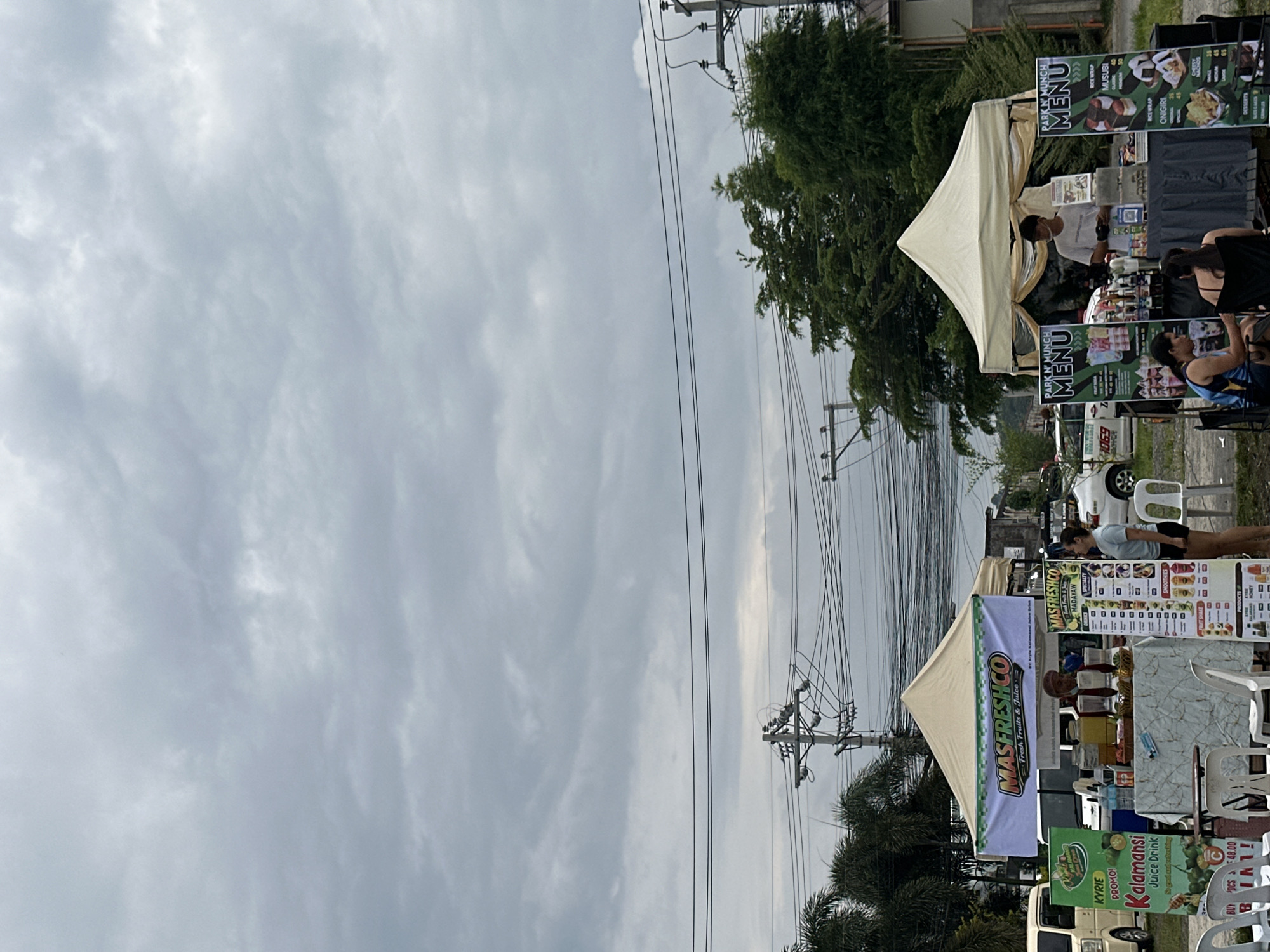This image captures an outdoor street food market scene, likely in Davao City, Philippines, under an overcast sky. The setting appears to be along a roadside, with electrical wires crisscrossing overhead and green trees bordering the area.
Two distinct vendor stalls are prominent. On the right side of the image, a light-colored tent belongs to "PARK N' MUNCH." Banners display their "MENU," featuring items like "RICE WRAP," "MUSUBI," "ONIGIRI," and "DESSERTS," with "40" visible as a price point. A male vendor stands within the tent, attending to customers. Several people are visible around this stall, some seated at white plastic tables and chairs, while others stand, suggesting ordering or waiting.
To the left, another tent houses "MASFRESHCO Fresh Fruits & Juice." Their signage includes "MASFRESHCO MADAYAW SPECIALTY" and a "KYRIE PROMO!" for "Kalamansi Juice Drink," described as "So good and refreshing." Other signs mention "SMOOTHIES" and "BUY DCS 1 48.00." A female vendor is seen behind a counter, facing customers. A light-colored vehicle is partially visible near this stall.
The activity depicted is that of a bustling street market, with vendors selling food and drinks and customers engaging with the stalls. The time of day appears to be daytime, possibly late afternoon, indicated by the diffused light from the cloudy sky. The overall atmosphere is casual and community-focused.