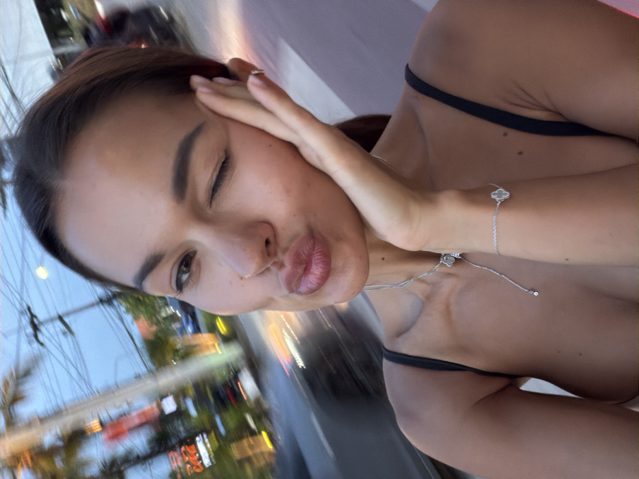 A selfie captures an adult female outdoors in Choeng Thale, Thailand, against a significantly blurred urban background, indicating movement or shallow depth of field. The subject, positioned centrally, has medium skin tone and dark hair, winking with her right eye while her left eye remains open. Her lips are pursed in a "kiss" expression, and her right hand is pressed against her right cheek. She wears a black thin-strapped top, a silver-colored necklace with a clover-shaped pendant, and a matching bracelet on her left wrist. The background features blurred vehicle lights, suggesting a street or road, alongside illuminated red and orange commercial signage, one appearing to display "CAFE." Palm trees and utility poles with numerous wires are also discernible against the dim sky, consistent with dusk or evening conditions.