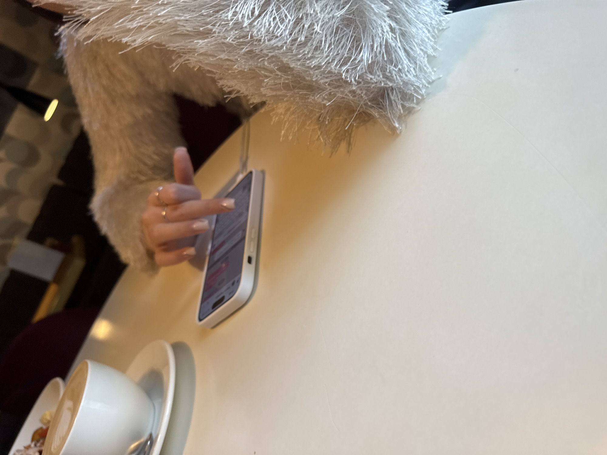 A close-up indoor shot depicts a light beige table with a smartphone, a cup of coffee, and a hand with rings on it. The fingers are tapping on the screen of the smartphone. The smartphone has a white case. The coffee cup appears to be white and partially obscures a saucer and a bowl of food. A fuzzy white garment is visible in the top left of the frame. The location and city cannot be determined from the image.