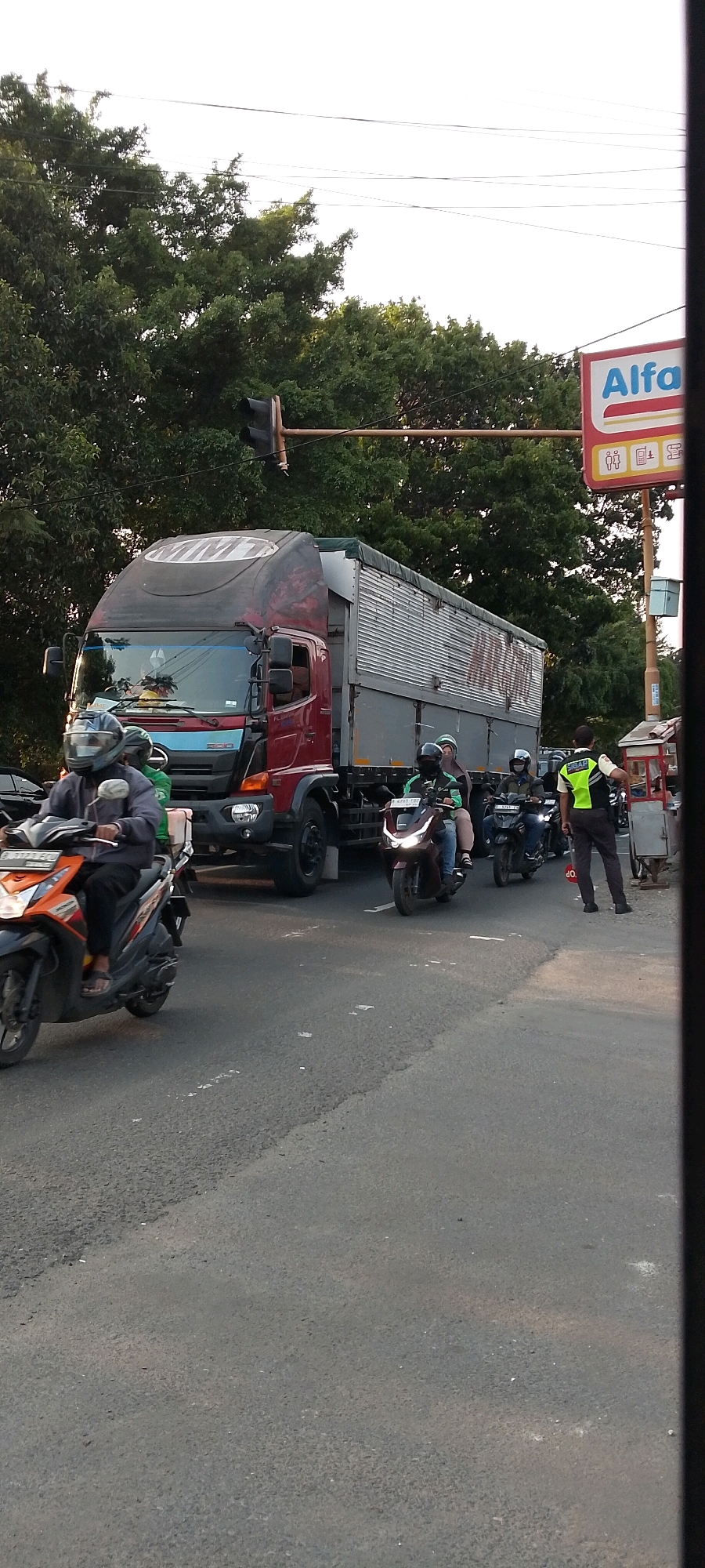 This is a forensic description of an image taken in Depok, Indonesia, at an intersection. A red and silver semi-trailer truck dominates the center of the frame, facing left. The truck's trailer is covered with a gray tarp. To the left of the truck, an orange and black scooter is stopped, ridden by a person wearing a blue jacket and a black helmet. Behind them, another scooter, dark red, is stopped, with two individuals on board wearing helmets. A person in a fluorescent yellow and green vest, who appears to be a traffic officer, stands to the right of the truck, holding a red stop sign. In the background, there are trees and a sign that reads "Alfa" with other logos. A traffic light is visible above and to the left of the truck. The road surface is asphalt, with some white lane markings. The sky is overcast.
