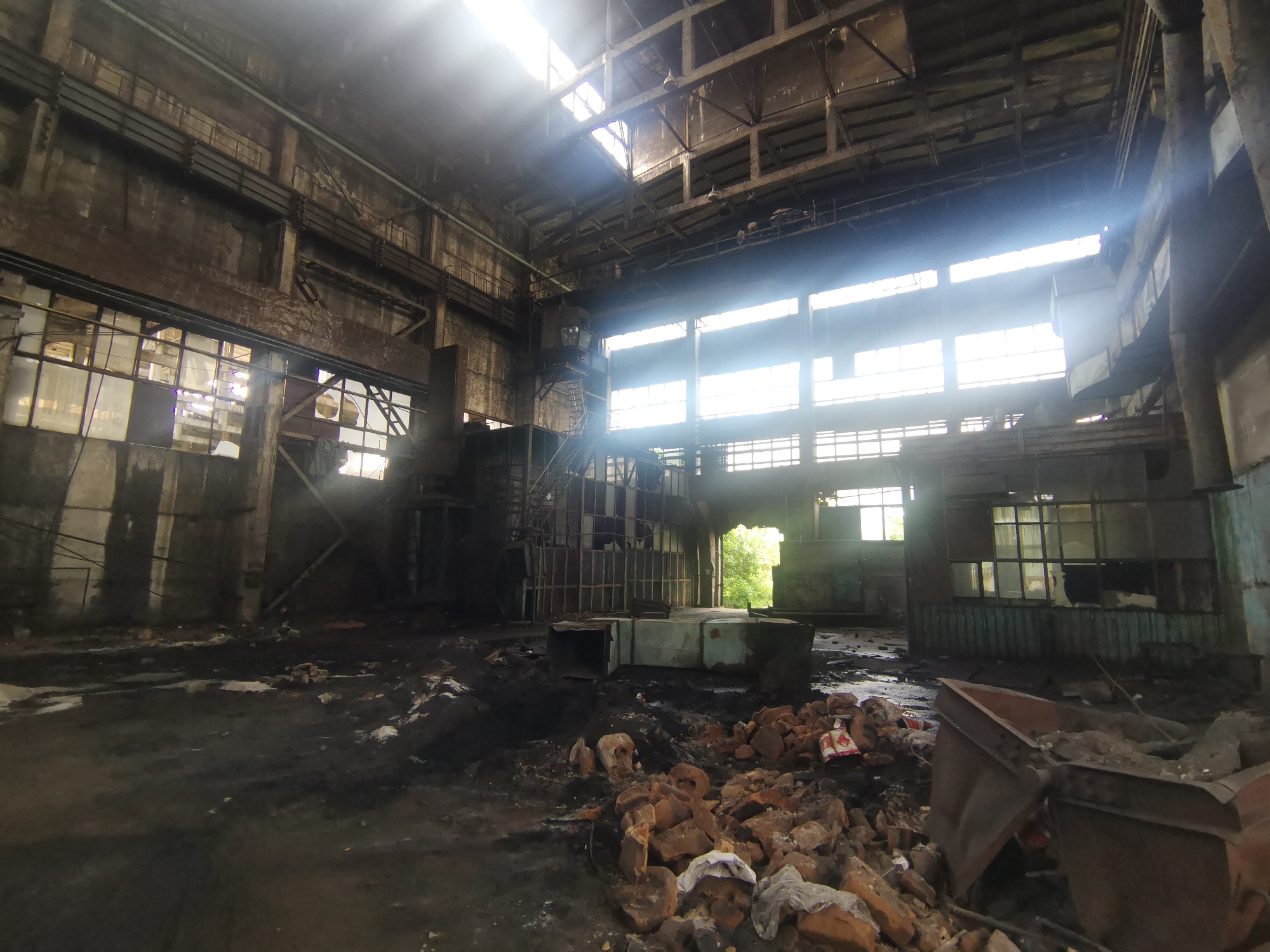 The image depicts the interior of an abandoned industrial building, likely located in or near Zhivkovo, Bulgaria. The space is characterized by decay and neglect. Natural light streams in through broken windows in the upper walls and roof, illuminating the debris-strewn floor. The floor is covered with dirt, dust, rubble, and scattered bricks. Various metal objects and structures are visible. The building appears to be a large, open warehouse or factory, with a high ceiling supported by metal beams and columns. There are no people visible in the image. There is a large opening that leads to a green landscape suggesting trees and other plant life, indicating the building’s exterior surroundings.