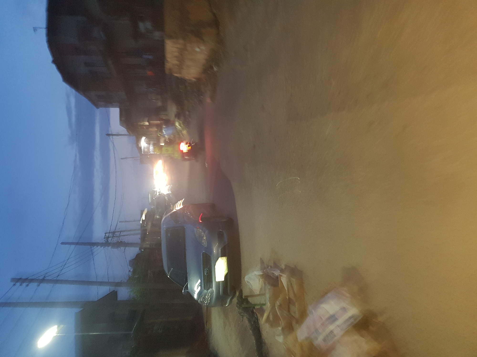 The image depicts a street scene in what appears to be Shangisha, Nigeria, during the evening or night. A silver car is parked on the right side of the road, partially obstructing the path. Debris, possibly trash bags, are scattered on the ground near the car. In the distance, there are lights from other vehicles, including a motorcycle. The buildings are visible on both sides of the street. There are telephone poles and a dimly lit street lamp on the left side of the frame. The sky appears to be a mix of blue and dark.