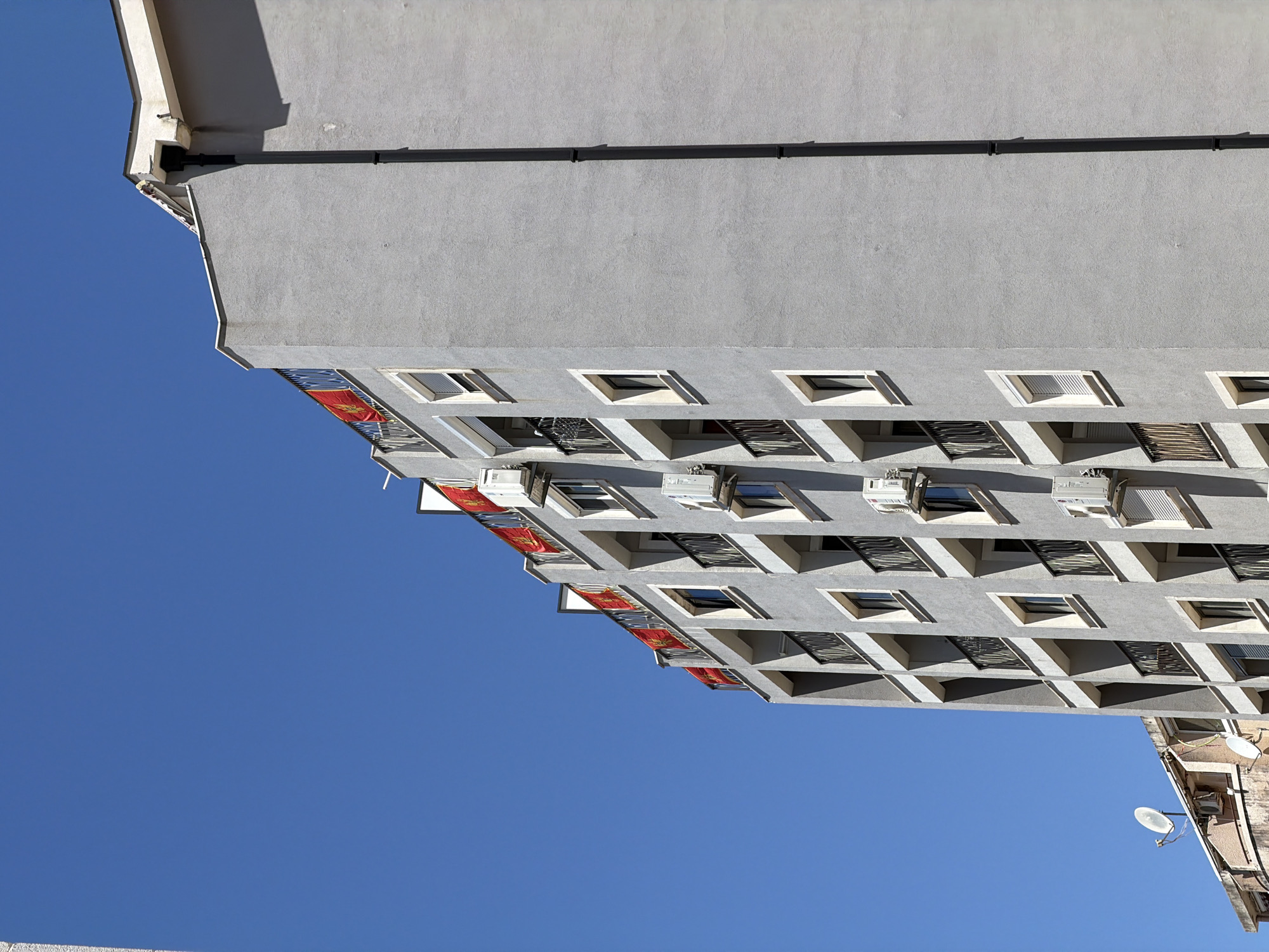 A low-angle shot captures an apartment building in Budva, Montenegro, against a cloudless blue sky. The building is pale gray with rows of windows and cantilevered balconies. Several air conditioning units are mounted below the windows. Some balconies display red flags or banners. A black pipe runs horizontally along the building's upper section. To the right, a neighboring building peeks into the frame, displaying a satellite dish on its exterior.