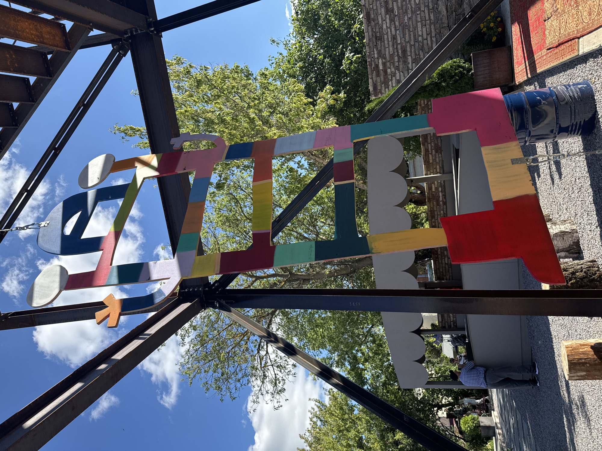 A bright, colorful, abstract metal sculpture resembling a ladder or abstract figure is suspended from a dark metal framework against a bright blue sky with white clouds. The sculpture is composed of various geometric shapes painted in a rainbow of colors, including red, orange, yellow, green, teal, light blue, pink, and purple. It appears to be an outdoor installation.
In the background, a man wearing a blue plaid shirt and gray pants is standing at a gray kiosk with a scalloped awning. Two people are visible behind the counter of the kiosk. To the right of the sculpture, a portion of a red patterned rug is visible on the ground, and a dark blue barrel is positioned beneath the lower part of the sculpture. The ground is covered in small gray gravel. Trees with green foliage are visible behind the sculpture and the kiosk. The overall environment suggests an outdoor public space. The location is identified as Yelets, Russia.