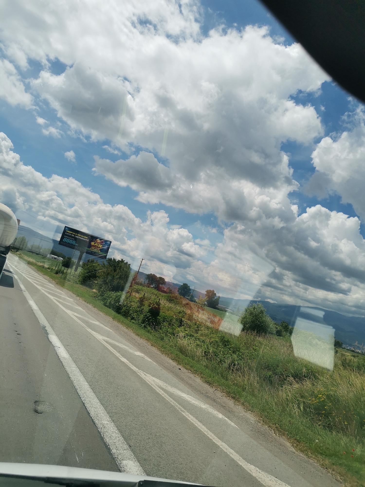 The image captures a scenic view from a vehicle moving along a highway, likely in or near the city of Krivina, Bulgaria. The foreground shows a paved road with white lane markings, indicating a clear path for traffic. To the right of the road, a lush green embankment covered with tall grass and scattered wildflowers slopes upwards. In the background, a billboard advertising a casino with the text "Оферти и печалби като в света" and the number "8888" is visible. Beyond the billboard, a line of trees forms a natural barrier, with rolling hills and distant mountains completing the landscape. The sky is a dynamic canvas of blue, dotted with large, fluffy white clouds, suggesting fair weather with some cloud cover. Reflections on the vehicle's window are also present, obscuring parts of the scene.