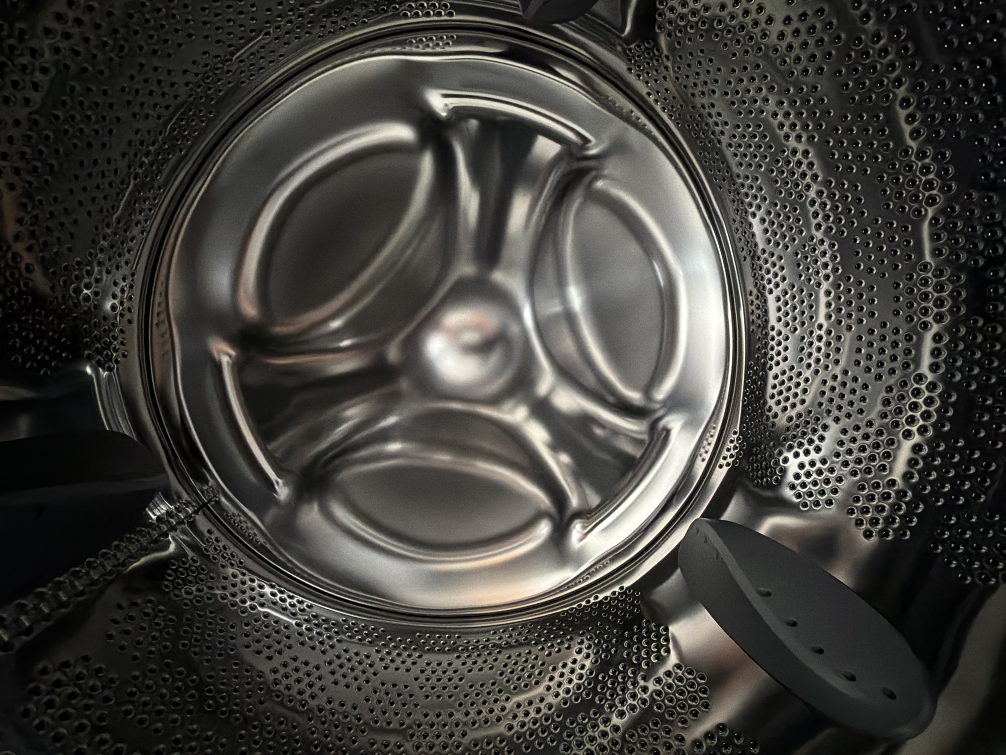 A close-up, interior view of a washing machine drum made of stainless steel. The drum is cylindrical with numerous small, evenly spaced perforations covering its inner surface. A central hub with three curved arms extends from the center of the back wall of the drum. A dark gray, elongated paddle or agitator is attached to the lower right side of the drum wall. The location in Ramat Gan, Israel is not determinable from the image.