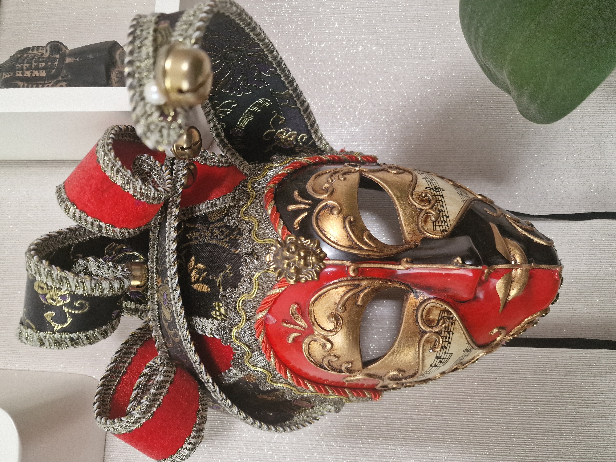 A Venetian-style mask hangs on a textured, light-colored wall. The mask is elaborately decorated with red, black, and gold accents. It features jester-like appendages adorned with gold trim, bells, and what appears to be a pearl. The face of the mask is split vertically, with one side predominantly red and the other black. Both sides are embellished with ornate gold scrollwork and musical notation. A small gold lion's head is visible on the red side of the mask, near the forehead. The background wall has a subtle vertical ribbed texture and a faint glitter effect. A small portion of a green, rounded object, possibly a plant leaf, is visible in the lower right corner of the image. The mask appears to be the sole subject in the foreground, with a blurred, dark figurine or artifact partially visible in the upper right quadrant, placed in front of a white framed rectangular object. The context of Nuremberg, Germany, is not discernible from the visual information provided.