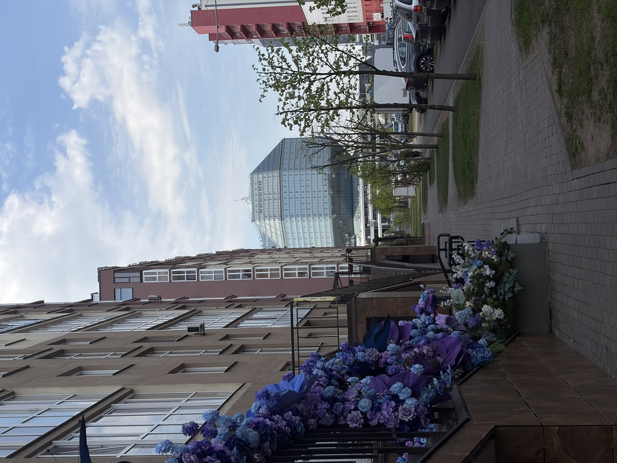 The image depicts a street scene in Minsk, Belarus. A brown building with many windows, adorned with purple and blue artificial flowers on a metal staircase, dominates the foreground. Beyond that is a glass office building, and a red and white building further up the street. Cars are parked along the curb, and small trees line the sidewalk. The sky is blue with some clouds. The image is angled with the scene rotated to the left.