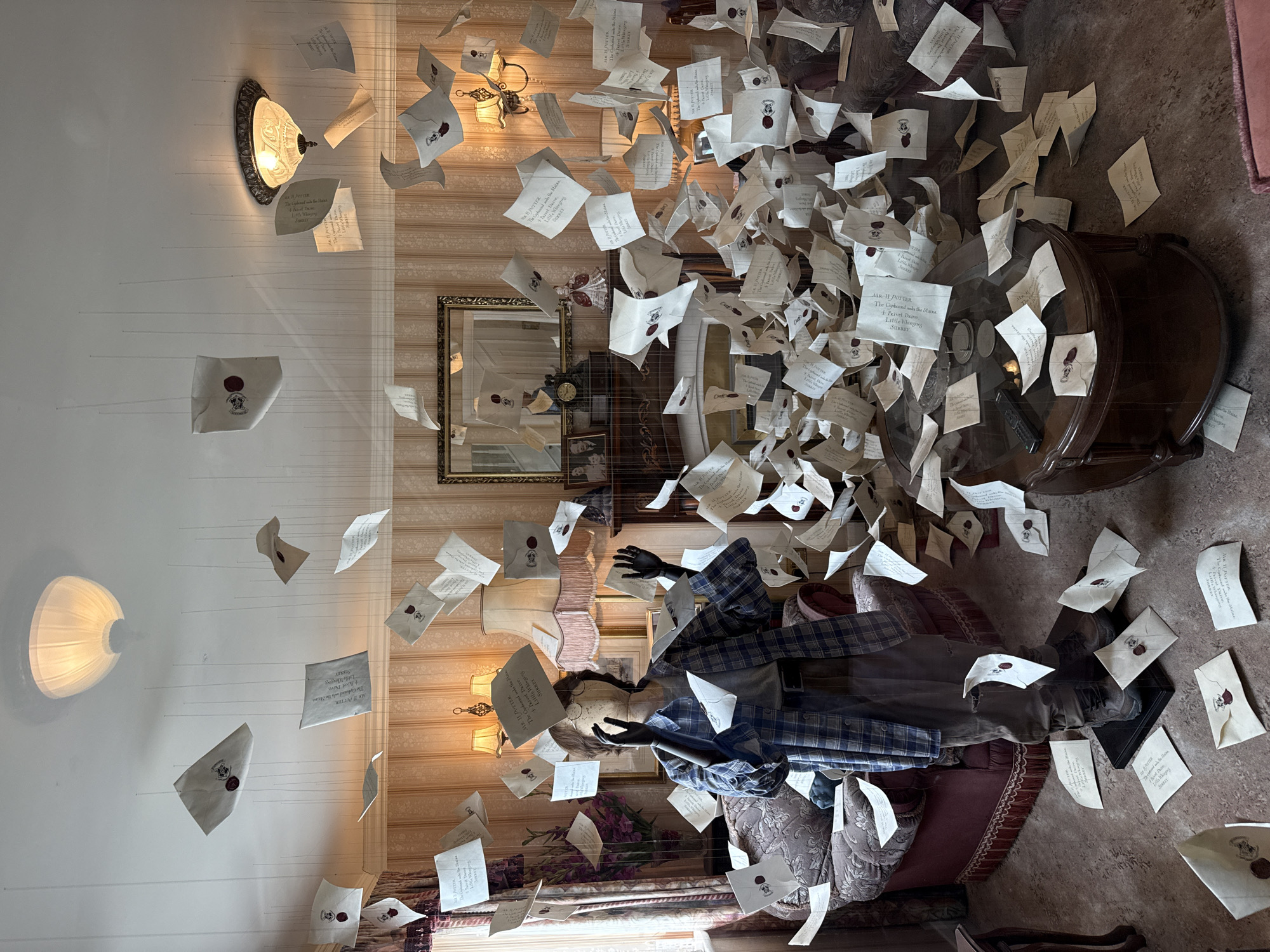 The image depicts an indoor scene, appearing to be a living room, filled with numerous letters suspended by thin wires from the ceiling. The letters are scattered throughout the room, some floating at different heights, with a dense cluster around a central coffee table and a mannequin. The mannequin, standing in the center, is dressed in casual clothing: a blue and white plaid shirt, grey t-shirt, dark trousers, and brown boots. It has dark gloves on its hands, which are raised as if in surprise or to catch the falling letters. The room's decor includes patterned wallpaper, lamps with fringed shades, a fireplace with a mirror above it, a sofa, and a wooden coffee table. The overall impression is a recreation of a scene from the Harry Potter series where Hogwarts letters are flooding into the Dursley's home. This scene is not located in the city of Three Rivers, United Kingdom.