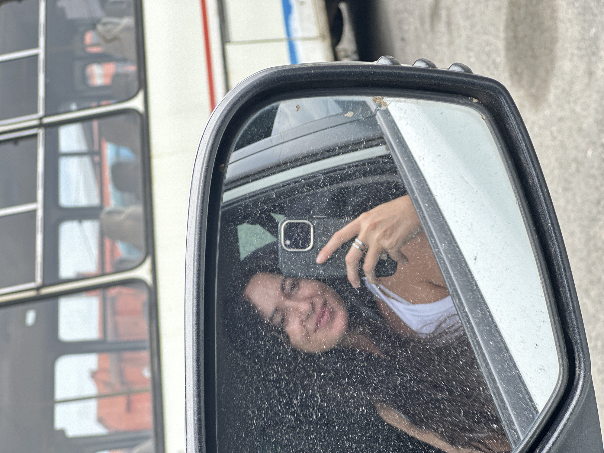 A woman with long dark hair is reflected in a car's side mirror, taking a selfie. She is wearing a white tank top and rings on her fingers. The mirror's surface is covered in water droplets, creating a distorted view of her face. In the background, a white bus with red and blue stripes is visible, its windows reflecting an orange building. The image appears to be taken in Slyudyanka, Russia, though this cannot be definitively confirmed from the visual evidence alone.