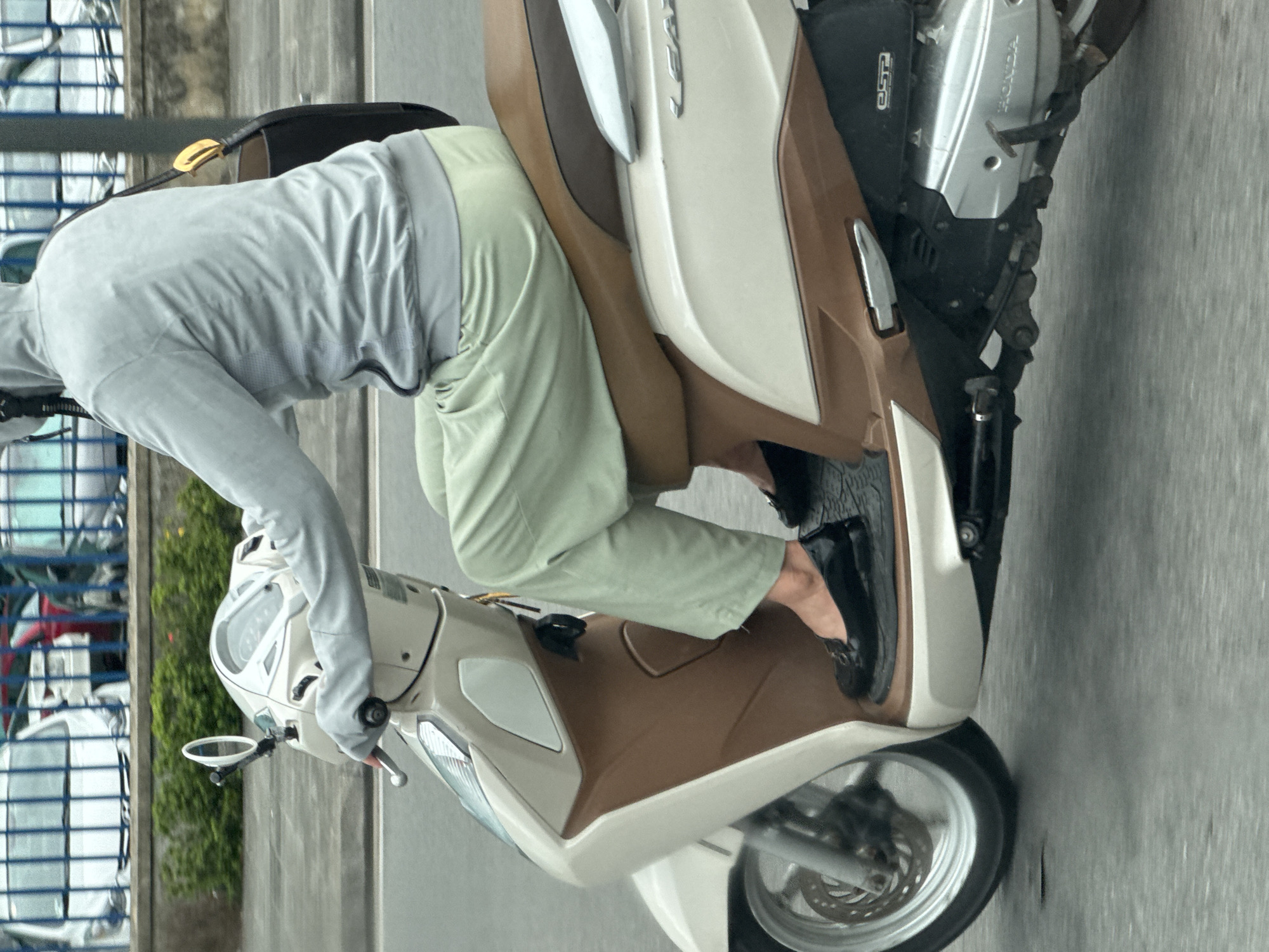 A person is riding a cream and brown scooter in Thuận Hóa Ward, Vietnam. The person is wearing a light gray hooded jacket and light green pants. They are also wearing black, flat shoes. A black bag with a gold buckle is slung over the person's shoulder. The scooter is on a paved road, and there are cars parked in the background, behind a low wall and some greenery. The image appears to be taken from a slightly elevated position, looking down at the rider and scooter.