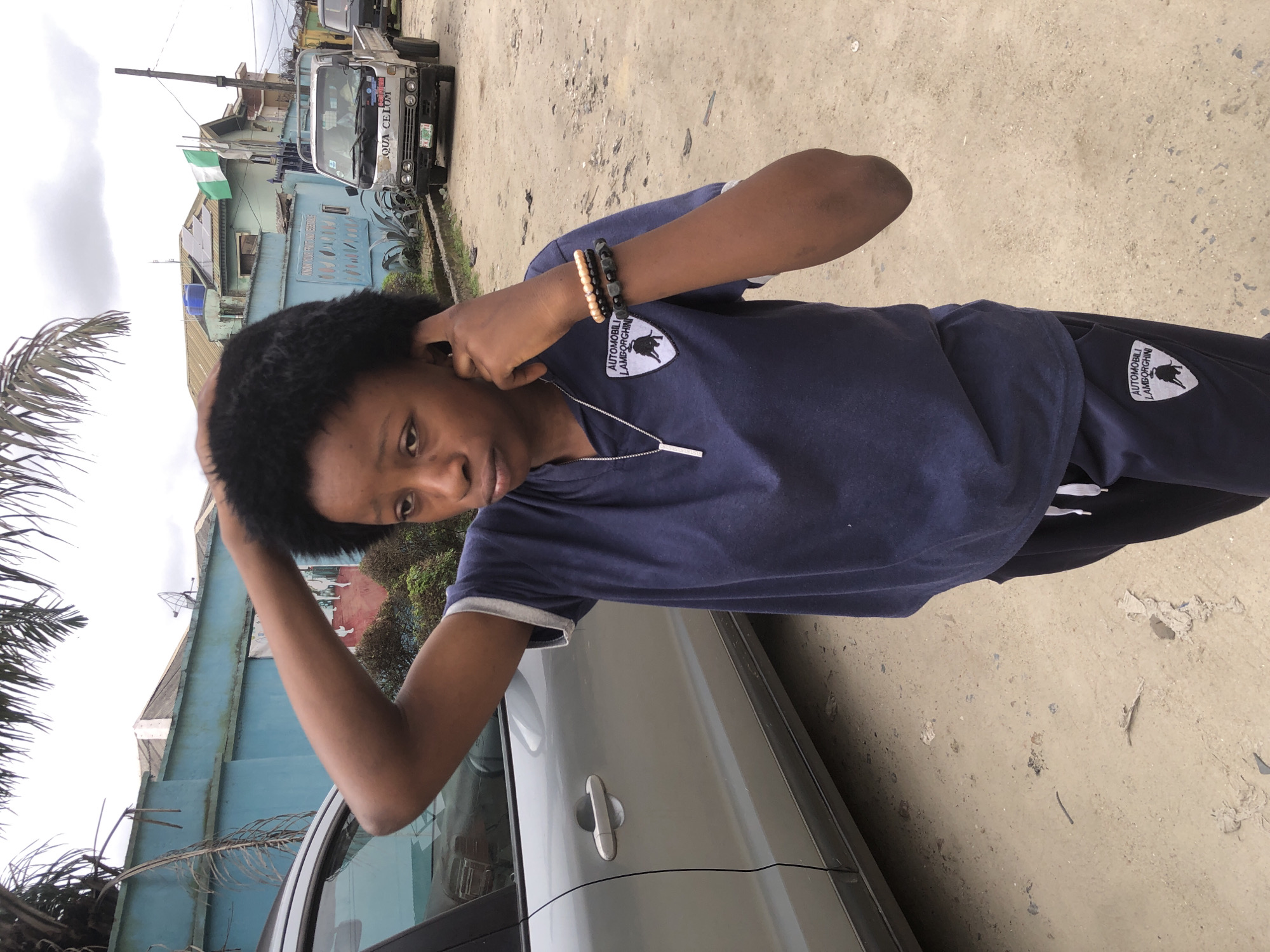 In Ado, Nigeria, a young woman stands on a dirt road next to a silver sedan. She wears a navy blue t-shirt with a white Lamborghini logo and navy blue pants with a matching logo. Her right hand is placed on her head, and her left hand is near her ear. She has two bracelets on her left wrist, one with beige beads and the other with dark beads. In the background, a light blue building with solar panels and a satellite dish is visible, along with a white truck and a Nigerian flag flying from a pole. The ground is covered in dirt and debris.