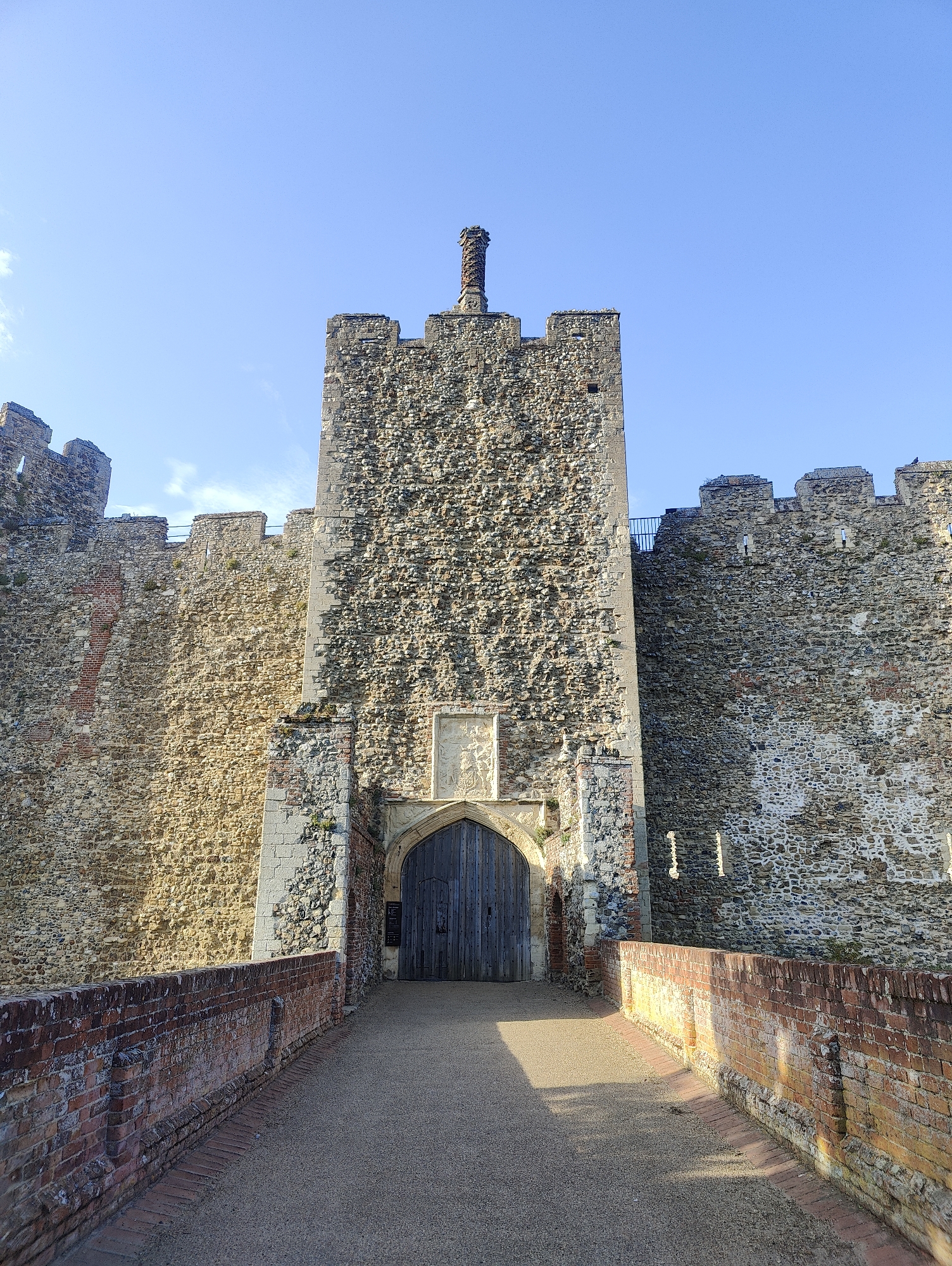 The image shows the main gatehouse of Framlingham Castle in East Suffolk, United Kingdom. The gatehouse is a tall, rectangular stone structure with a doorway at ground level and a smaller brick chimney atop the roof. The walls flanking the gatehouse are constructed of stone and brick, featuring crenellations along the top. A brick pathway leads to the gatehouse entrance. The sky is clear and blue. The time of day appears to be midday or early afternoon, judging by the shadows. No people are visible in the scene.