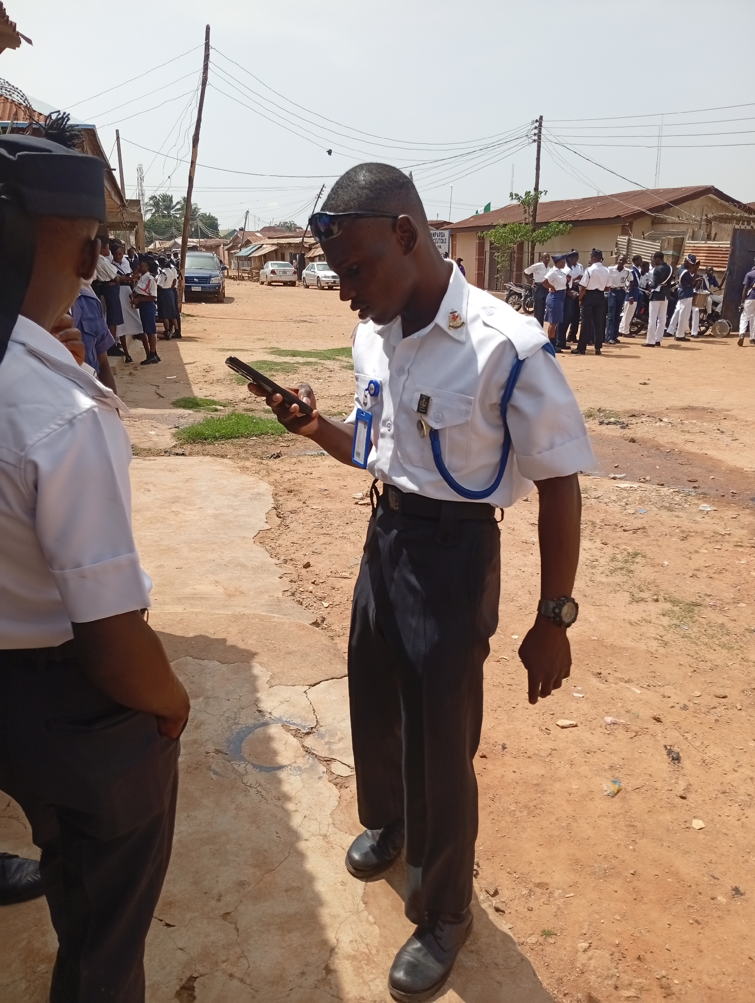 The image shows two people in uniform standing in an outdoor location, likely in the city of Unknown, Unknown. The person in the foreground is looking at their phone, and has a blue strap on their left shoulder. The other person is standing with their back to the viewer. In the background, there are other people in similar uniforms standing near some buildings, and several cars are parked on the street. The ground appears to be dirt.