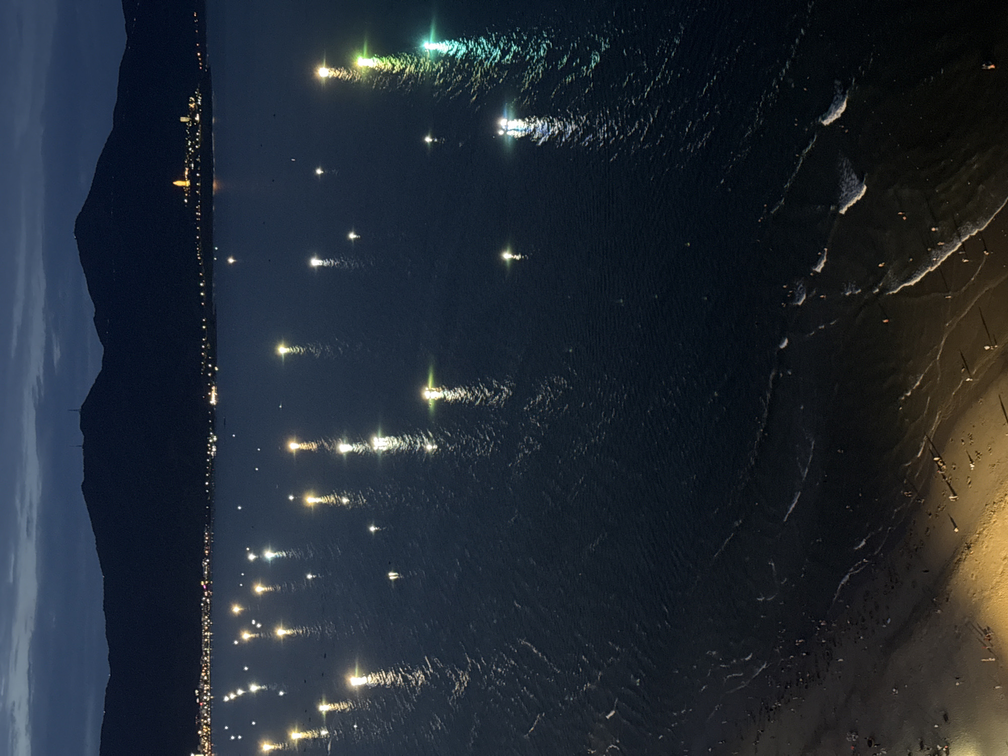 The image, taken at dusk or night, presents a high-angle view of a coastal scene in Đà Nẵng, Vietnam. On the left, a large, dark mountain range dominates the silhouette against a deep blue, twilight sky. Along the base of this mountain, a vibrant line of city lights stretches into the distance, illuminating the coastline. One particularly prominent, bright yellow light structure, possibly a temple or landmark, stands out among the urban glow.
The central part of the image is dominated by the dark, rippled surface of the water, likely the sea or a bay. Numerous bright, elongated reflections of the city lights shimmer across the water, distorted by the gentle waves. These reflections exhibit a range of colors, from warm yellows and whites to cool greens and blues, some showing a shimmering, almost iridescent quality. Several distinct, brighter light sources, possibly from boats or buoys, are also reflected as individual points of light further out in the water.
To the far right, a sandy beach is dimly lit, with gentle white-capped waves breaking along the shore. Small, indistinct figures, appearing as dark silhouettes, are scattered across the beach, suggesting people enjoying the evening by the sea. Some objects that could be beach umbrellas or structures are also faintly visible. The overall impression is one of a tranquil yet active coastal city at night, viewed from an elevated perspective. No visible text is present in the image.