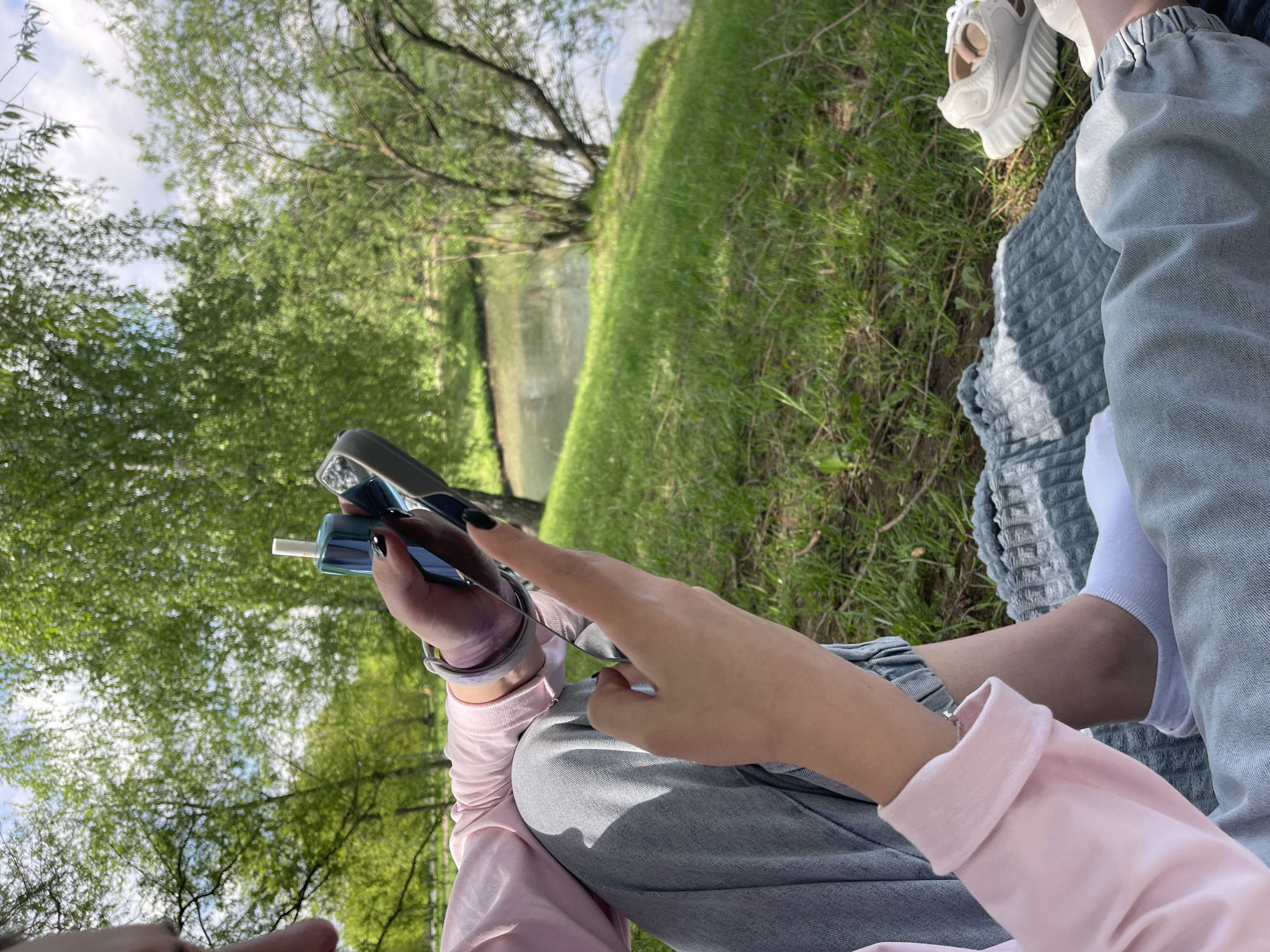 The image shows a person sitting outdoors, possibly in a park. The person is wearing gray jeans, white socks, a pink shirt, and white shoes. They have a device in their hand, possibly an electronic cigarette or similar item. The background shows green grass, trees, and a body of water. The location could be a park in Moscow, Russia, given the typical landscape features and vegetation found in the city.