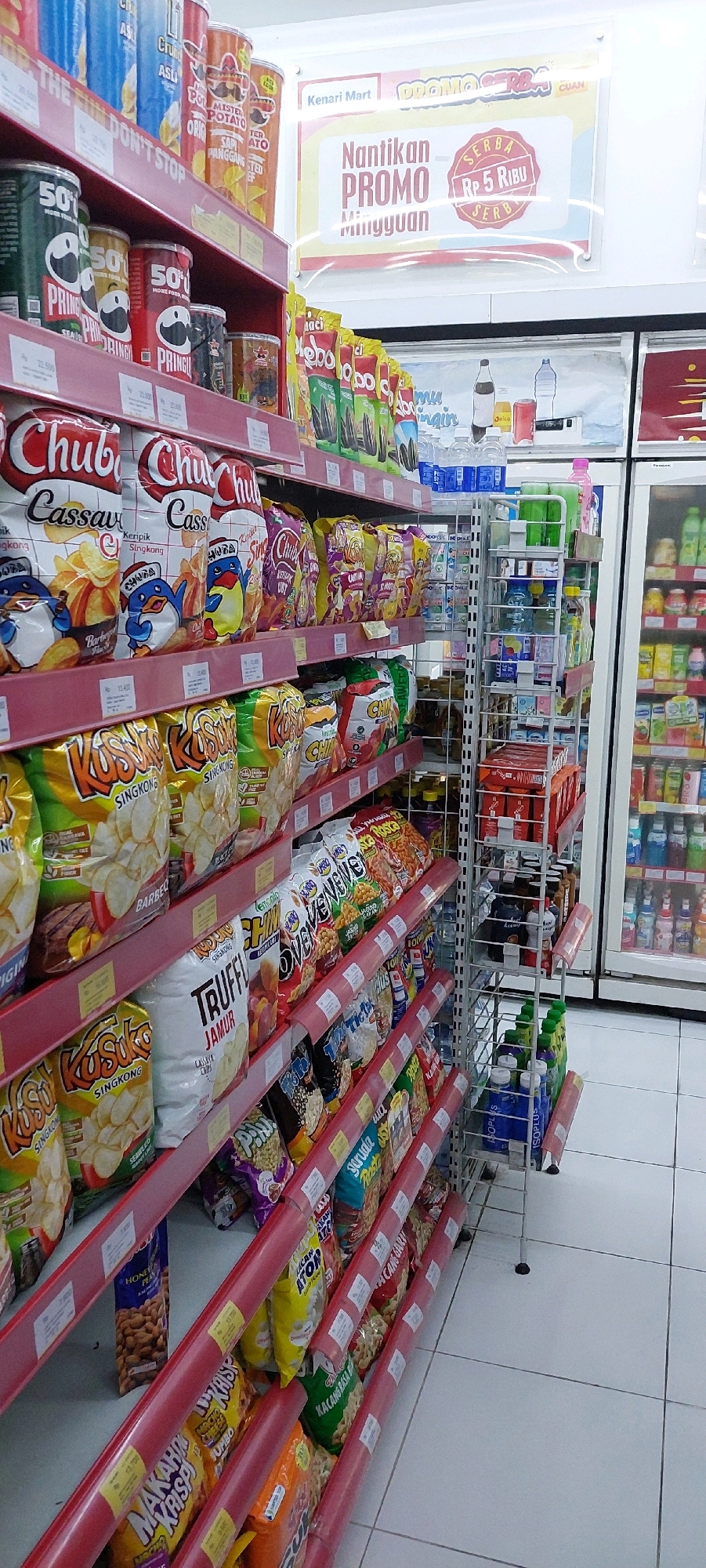 The image shows a brightly lit interior of a convenience store, likely a "Kenari Mart" as indicated by signage at the top. Shelves are filled with various packaged snacks, predominantly chips and crackers, in colorful bags and canisters. The shelves are made of red metal and are stocked from floor to ceiling. On the left, there are tall canisters of Pringles potato chips. In the middle, bags of "Chuba Cassava" chips are visible, some featuring cartoon penguins. Further right, there are rows of various bagged snacks, including "Kusuka Singkong" and "Truffle Jamur." Towards the back, a display rack holds bottles of drinks and smaller packaged goods. To the far right, a glass-doored refrigerator displays rows of bottled beverages, primarily water and flavored drinks. A promotional banner hangs above the shelves in the background, announcing "Nantikan PROMO Mingguan" (Anticipate Weekly PROMO) with a price indication of "Rp 5 Ribu" (Rp 5 Thousand). The flooring appears to be white tiles. No people are visible in the scene. The lighting is even and bright, suggesting an indoor retail environment.
