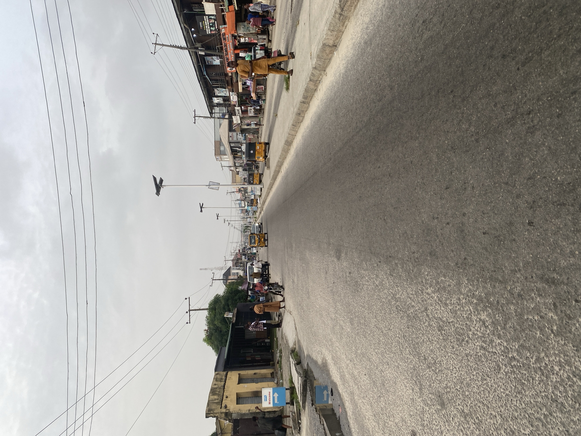 The image depicts a street in Ado, Nigeria, under an overcast sky. The road extends into the distance, lined with buildings and utility poles. Motorbikes and auto rickshaws (tuk-tuks) are present on the road. People are seen walking on the sidewalk and standing near buildings. A blue and white "Welcome" sign is visible near the bottom of the image. The buildings along the street appear to be a mix of commercial and residential structures. Overhead power lines traverse the scene.