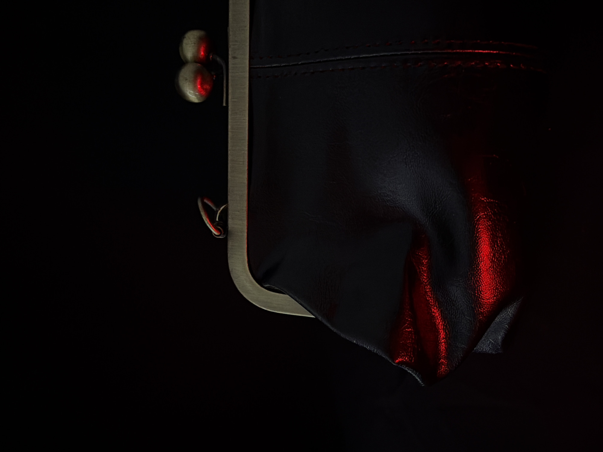 A close-up, vertical, high-angle shot shows a black coin purse with a metal clasp against a black background. The coin purse is angled with its opening facing left, and it is illuminated by a red light that casts a strong reflection on the lower left side of the purse. The metal clasp is a dull gold color and has two round, shiny balls on the right side. The purse appears to be made of leather and has visible stitching down the right side. The lighting is dim and dramatic, with the red light being the only source of color. The image is crisp and in focus, highlighting the texture of the leather and the metallic sheen of the clasp. There are no people or other objects visible in the scene.