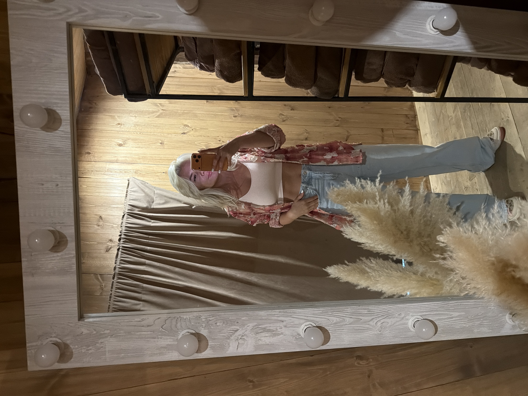 A full-length mirror shows a woman taking a selfie indoors. The mirror is framed with a light-colored, possibly wooden frame featuring bulb lights. The woman has long blonde hair and is wearing light-wash jeans, a white top, and a floral patterned shirt. She is holding a smartphone to take the photo. In the reflection, behind the woman, there is a tan curtain. Above her, there is a wooden surface with rolled towels placed on a black metal shelf. To the right of the woman, there is a bouquet of fluffy, light-colored pampas grass. The walls of the room appear to be made of wood.