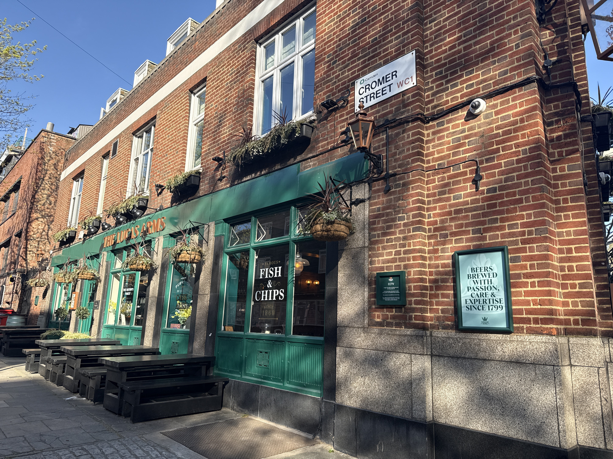 The image depicts the exterior of a traditional British pub called "The Lucas Arms", located on Cromer Street, WC1, in London, United Kingdom. The pub is a red-brick building with green-painted window frames and awnings. Hanging baskets adorn the facade.
There are outdoor seating areas with dark wooden tables and benches. The windows feature lettering indicating "Fish & Chips" and "Since 1860", indicating that the pub offers this popular dish. Signs on the building indicate "CCTV" surveillance and advertise "Beers brewed with passion, care & expertise since 1799".
The street sign "Cromer Street WC1" is visible. The photograph is taken during daylight, with a clear blue sky visible in the background. No people are visible in the frame.