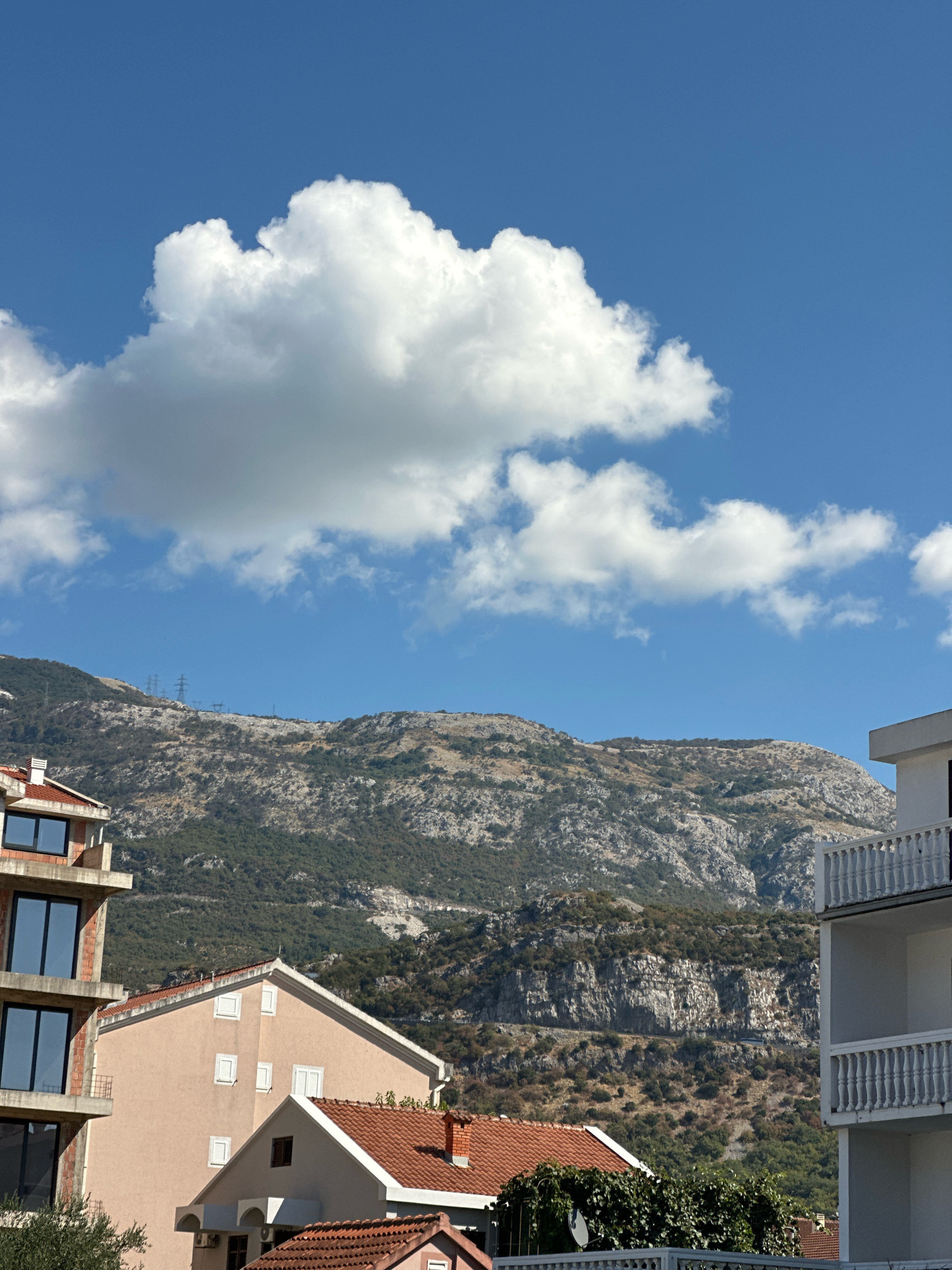 The image, taken in Budva, Montenegro, presents a view of buildings against a backdrop of a rugged, vegetated mountain under a bright blue sky dotted with fluffy white clouds. The foreground features the partial facades of several residential buildings. On the left, a modern structure with multiple stories and large, dark-framed windows is under construction, revealing its brick and concrete framework. Adjacent to it, a lighter-colored building with multiple windows and a red-tiled roof is visible. To the right, a white building with a balcony and ornate railing is present. The midground and background are dominated by a steep mountain slope covered in scrub and trees, with rocky outcrops. Power lines and pylons can be seen ascending the mountain in the distance, suggesting the presence of infrastructure. The overall scene conveys a sense of a coastal town nestled against a mountainous landscape on a clear day.