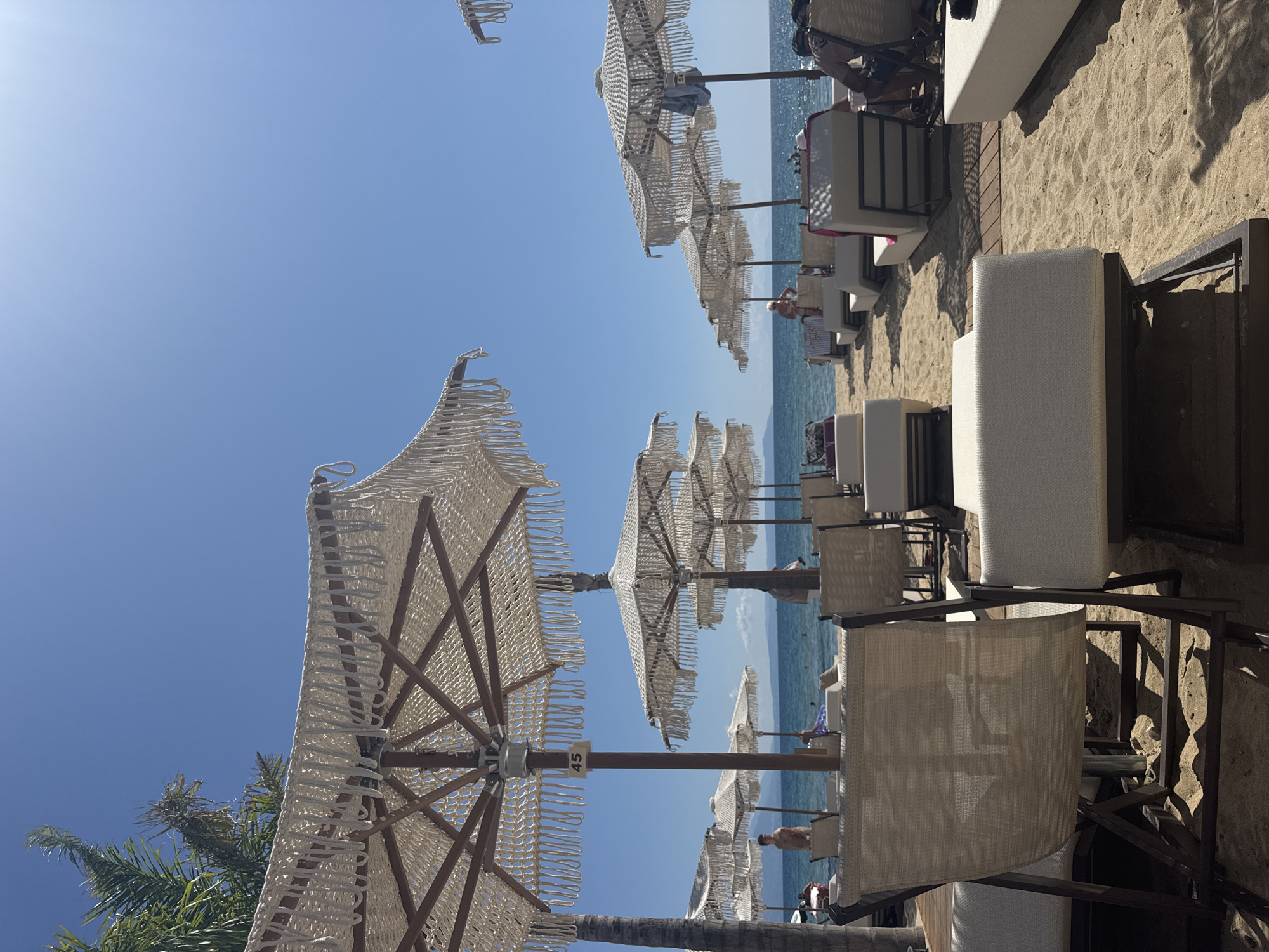 This is a bright, sunny photograph taken on a sandy beach in Stavros, Greece, likely on a resort. In the foreground, there are several empty, plush white lounge chairs and some empty beige beach chairs. Behind these, a row of large, white beach umbrellas with decorative fringe are set up on the sand. Some of these umbrellas are numbered, with one clearly showing the number "45". A few people are visible in the background, some relaxing on lounge chairs under umbrellas, and others walking or swimming in the clear blue water of the Aegean Sea. The sea stretches to the horizon where a hazy landmass is visible. The sky is a clear, bright blue with no clouds. Palm trees are present on the left side of the image, contributing to the tropical atmosphere. The overall scene depicts a serene and idyllic beach resort setting.