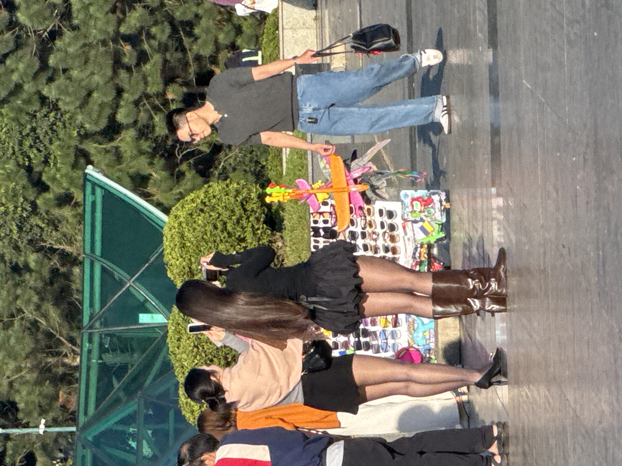 The image captures a street scene in Da Lat, Vietnam. In the foreground, a person wearing a black outfit with a puffy skirt and tall brown boots is posing for a photo, holding a camera. To their left, another person in a light pink top and black skirt is also taking a picture with their phone. A man in a dark grey short-sleeved shirt and blue jeans walks by, carrying a black bag. Behind them, a stall displays various items, possibly sunglasses. In the background, lush green trees and a large, modern glass structure suggest an urban or park-like setting. The ground appears to be paved, with dappled sunlight and shadows indicating an outdoor, daytime environment.