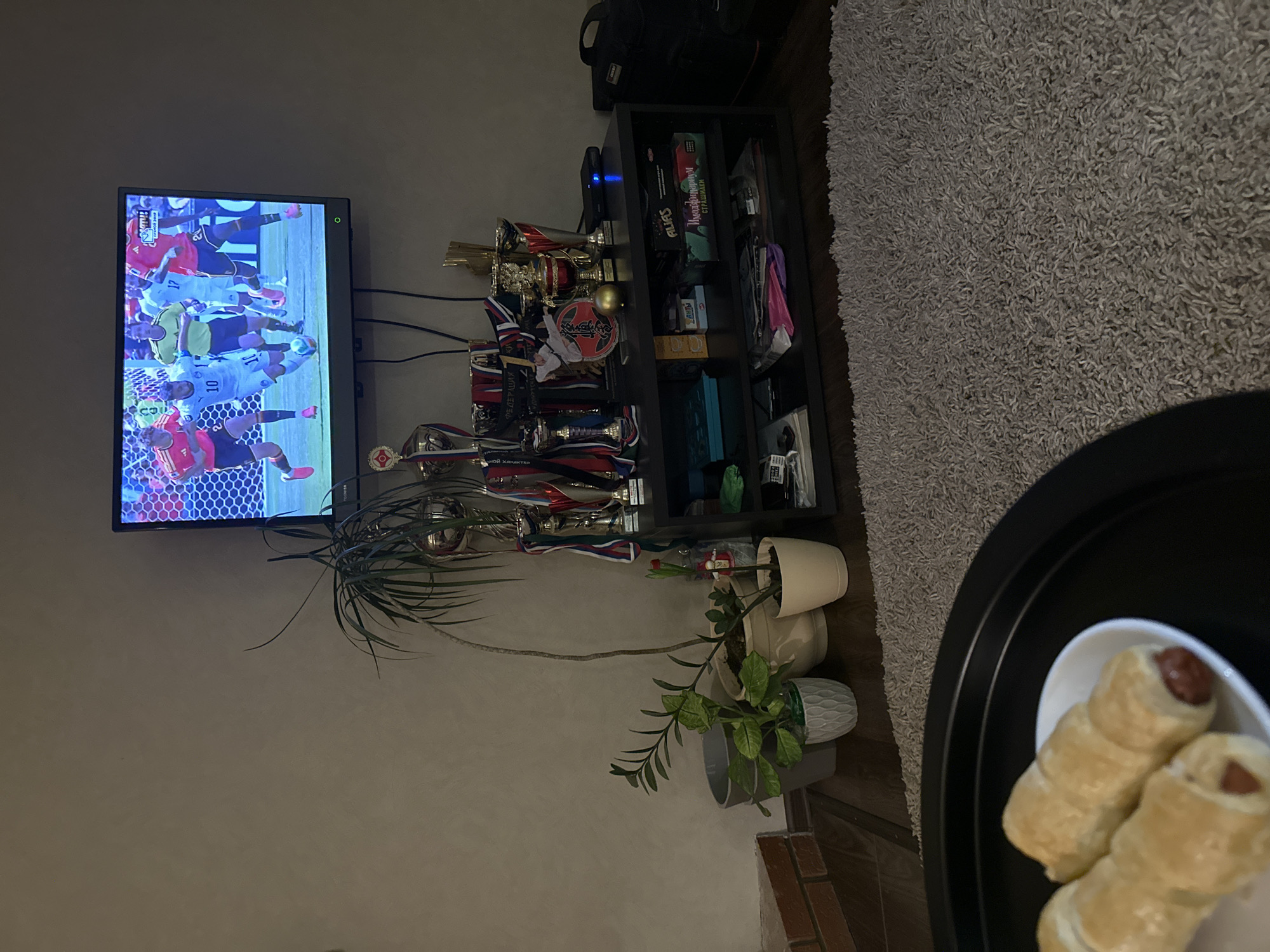 The image depicts a dimly lit living room with a flat-screen television mounted on the wall, displaying a soccer match. In front of the television, a black entertainment unit holds several trophies and decorative items. To the left of the unit, there are three potted plants. In the foreground, a black tray holds a white plate with two pastry-wrapped sausages. The room has a grey carpet and a partial view of a brick fireplace on the left. The image does not provide any specific indicators of its location within Chaikovsky, Russia.