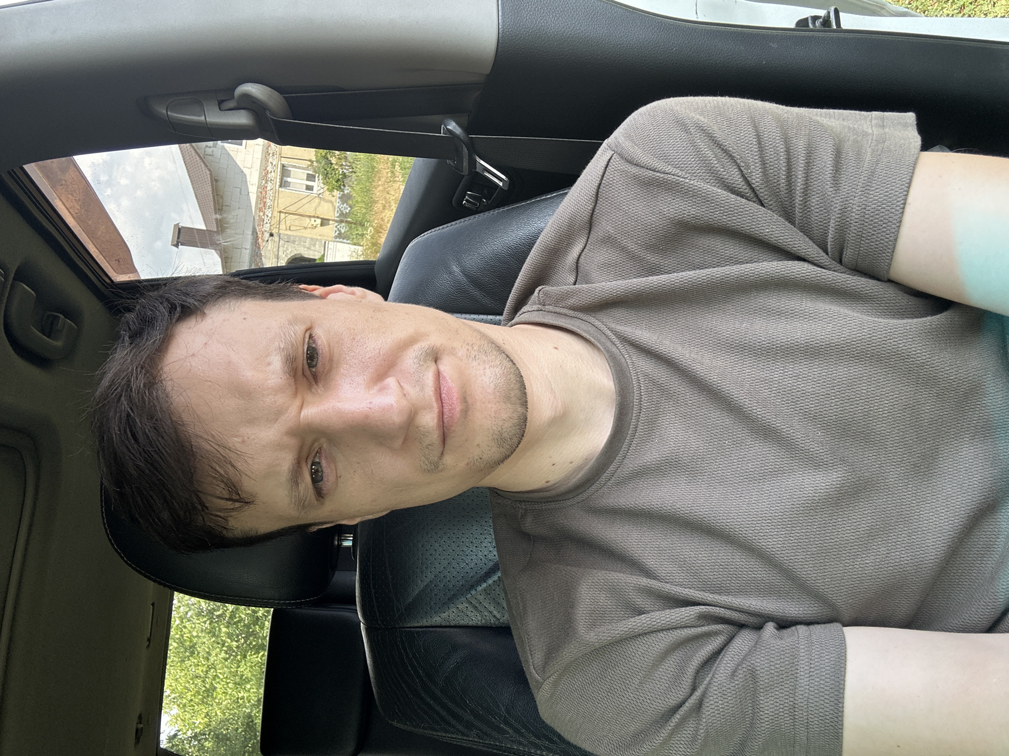 A middle-aged man with short, dark hair and a light beard is sitting in the driver's seat of a car, looking at the camera. He is wearing a brown t-shirt. The interior of the car is visible, including the black leather seat and a seatbelt. Outside the car, a green tree and a house with a tile roof can be seen. The location is the city of Makiv, Ukraine.