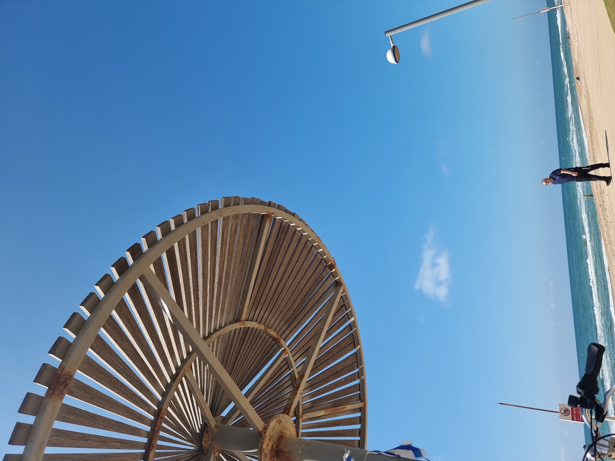 A low-angle shot depicts a partial view of a round, sun-shading structure comprised of wooden slats and metal framework, under a bright blue sky with a few clouds. At the far right, a beach is visible with a man walking along the shoreline. In the distance, a lamppost extends into the sky. In the foreground on the lower-right corner, a bicycle handlebar and part of a sign are visible. The scene appears to be taken at a beach in Tel Aviv, Israel, which is identifiable by the style of the sun-shading structure and beach.