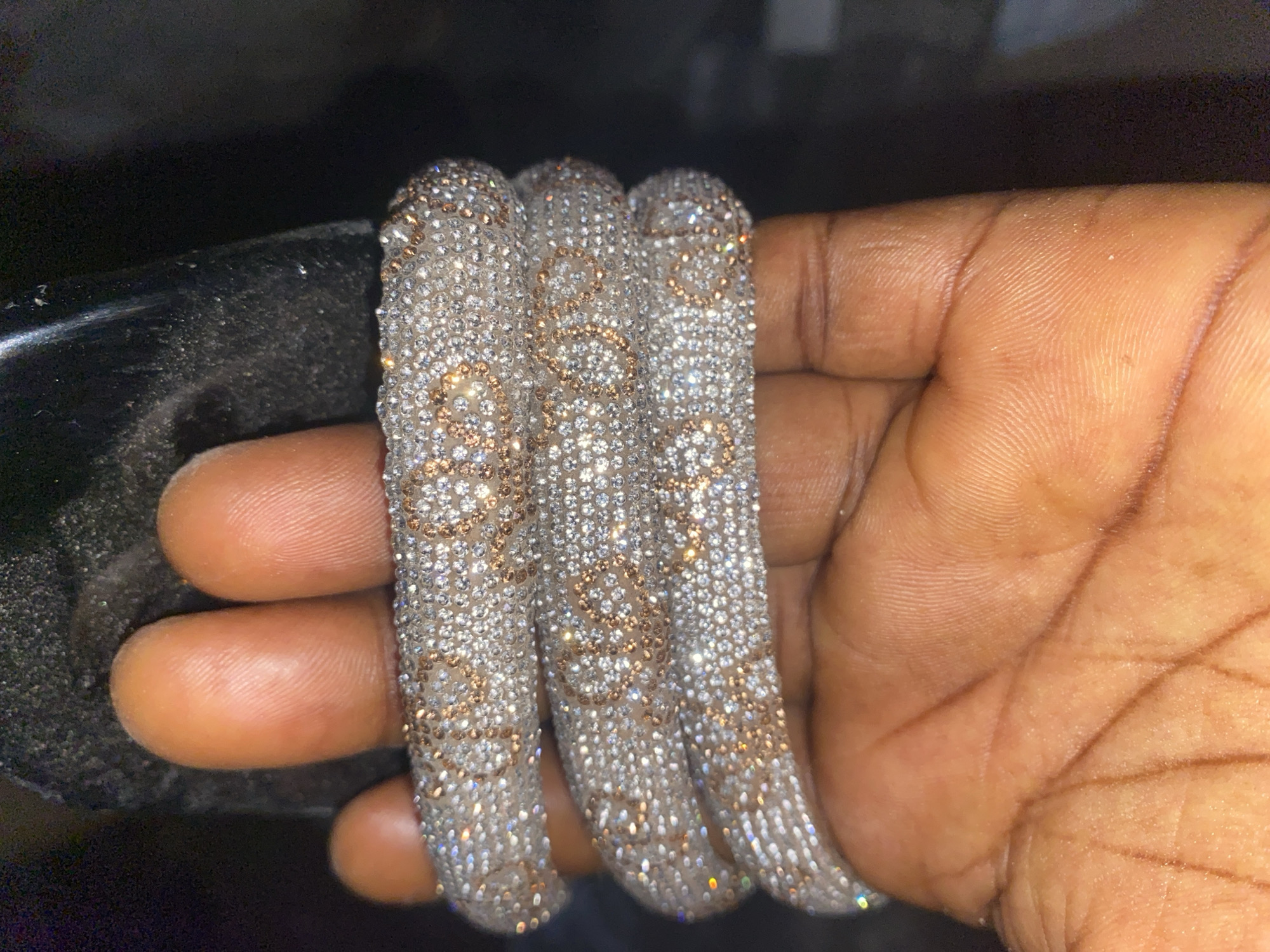 The image shows a close-up of a hand holding three stacked diamond-encrusted bracelets. The bracelets are thick and appear to be made of white gold with rose gold accents forming an intricate pattern. The diamonds are small and densely set, giving the bracelets a sparkling, luxurious appearance. The hand holding the bracelets has dark skin with visible fingerprints and palm lines. The background is dark and out of focus, suggesting an indoor setting or a low-light environment. There is no identifiable location within the city of Jauro Yinu, Nigeria, as the image is a close-up and lacks any contextual landmarks. The image focuses solely on the hand and the jewelry.