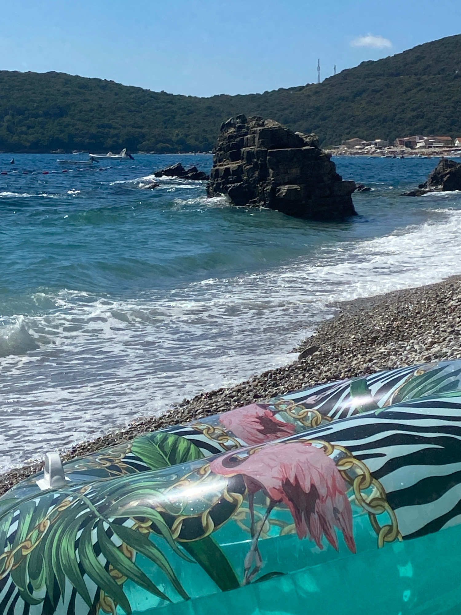 In Prijevor, Montenegro, on a sunny day, two inflatable pool floats are positioned on a pebble beach. The floats are decorated with a tropical print featuring pink flamingos, green palm leaves, and gold chains against a background of zebra stripes. The sea is blue with gentle waves breaking on the shore. In the background, a rocky island formation rises from the water, and a densely forested hillside with some buildings is visible. The sky is clear and blue.