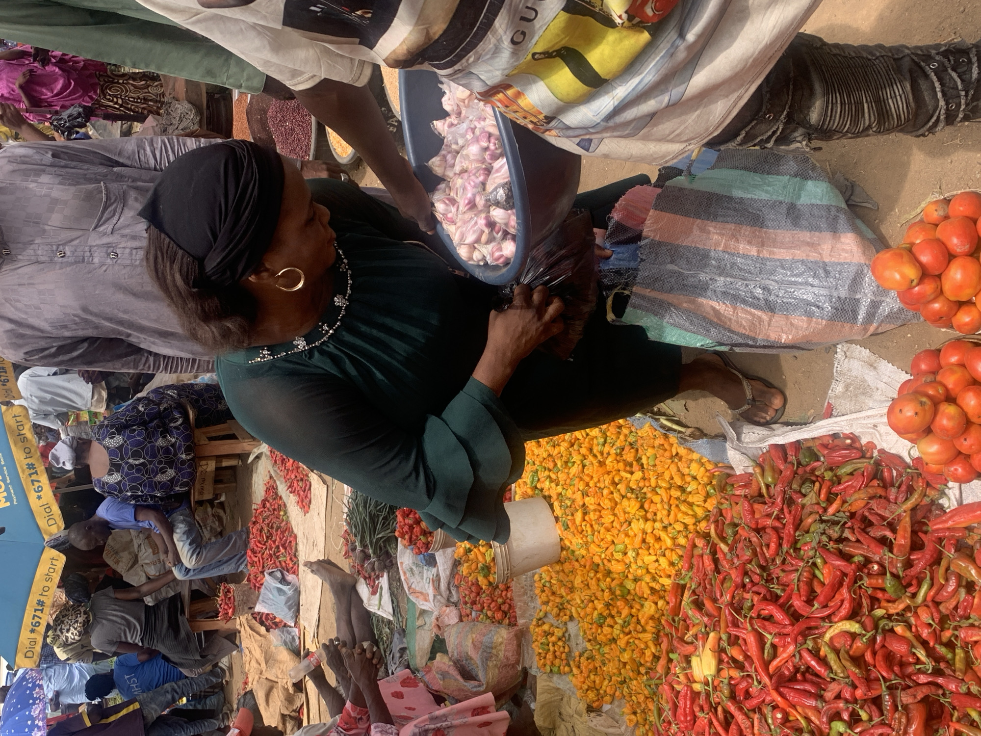 The image depicts a bustling marketplace scene likely in Jos, Nigeria. The main subject is a woman wearing a dark green dress and a black head covering. She is engaged in purchasing garlic from a vendor who is holding a blue container filled with garlic. The woman is holding a black plastic bag, presumably to receive the garlic. Surrounding the woman and vendor are various produce items. There are piles of tomatoes, red chili peppers, and yellow habanero peppers. Other vendors and customers are visible in the background, adding to the busy atmosphere of the market. The ground is light-colored. Other goods are also visible, indicating a diverse array of products being sold. An advertisement can be seen on the left.