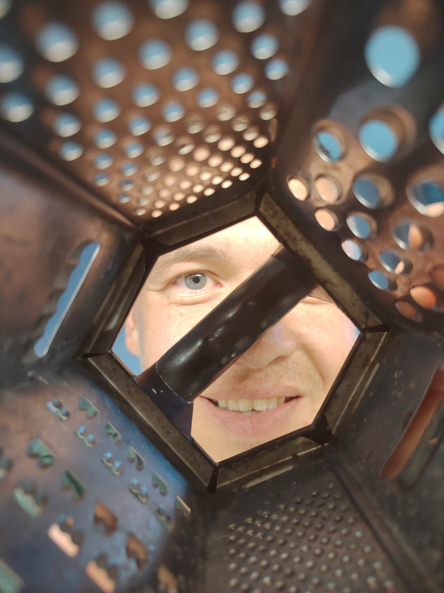 A playful, close-up shot captures a person looking directly through the central opening of a dark, metallic box grater. The grater, likely a kitchen tool, occupies most of the foreground and midground, its four sides displaying various grating surfaces with different sized and shaped holes, including circular, tear-drop, and sharp-edged perforations. A prominent black bar or handle runs horizontally across the hexagonal opening through which the person's face is seen.
The subject, appearing to be a man, is smiling broadly, revealing white teeth, and has a clear blue eye visible. His expression conveys amusement and engagement. Parts of his forehead, cheek, and nose are also visible, framed by the grater's structure.
Through the multiple holes of the grater and the central opening, a bright, clear blue sky is visible in the background, suggesting an outdoor setting during daytime with good weather. The lighting is natural and bright. The image does not contain any visible text. This photograph was taken in Chaikovsky, Russia.