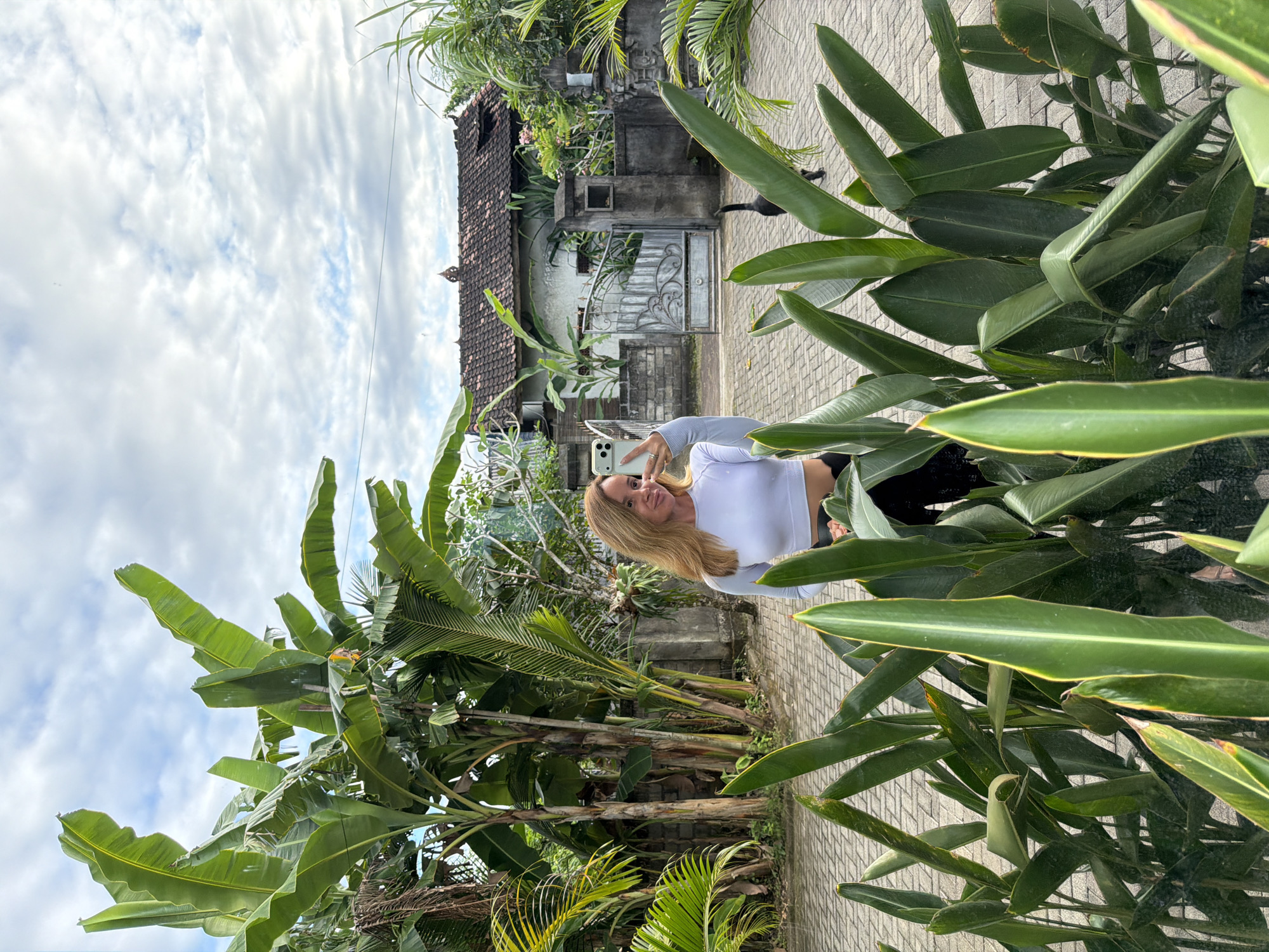 A blonde woman takes a selfie outdoors in Kuta Selatan, Indonesia. She wears a white long-sleeved shirt and black pants, holding her phone up with her right hand. Behind her is a weathered building with a tiled roof and a light-colored gate. A black cat walks behind the gate. Tropical plants frame the scene. The ground is paved with light gray bricks. The sky is cloudy.