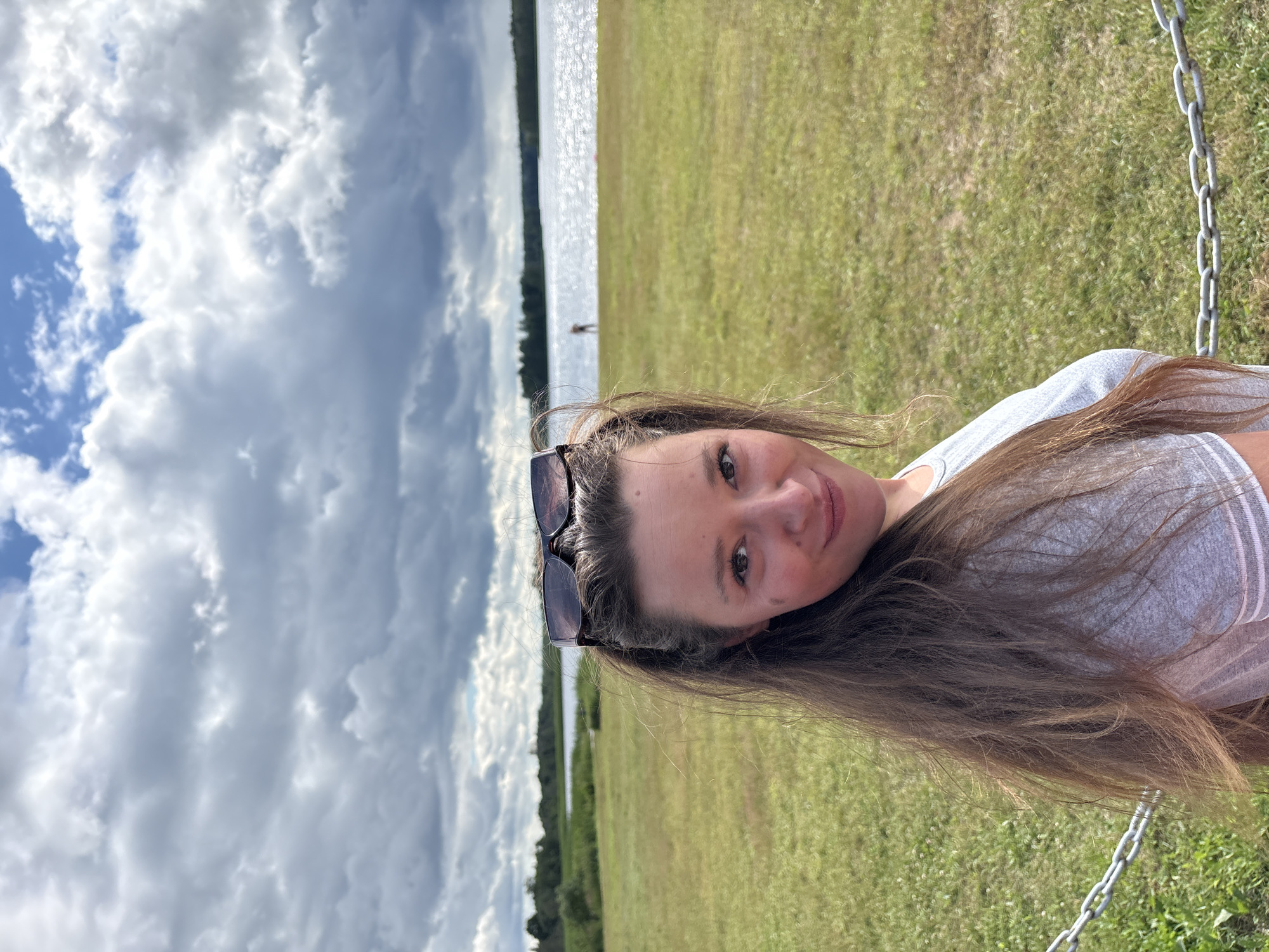 The image features a young woman posing for a selfie outdoors. She has long, brown hair and is wearing sunglasses on top of her head. She is looking at the camera with a relaxed expression. The background shows a body of water with a faint reflection of the sky, suggesting it is a sunny day. To the right of the water, there is a grassy field. In the distance, a lone figure can be seen near the water's edge. The sky is partly cloudy with dramatic cloud formations. A metal chain, likely part of a swing set, is visible in the lower right corner of the frame. The orientation of the image suggests it might have been taken in a vertical format. The scene appears to be a natural, possibly recreational area, consistent with the provided location context of Мазалаўскі сельскі Савет, Belarus.