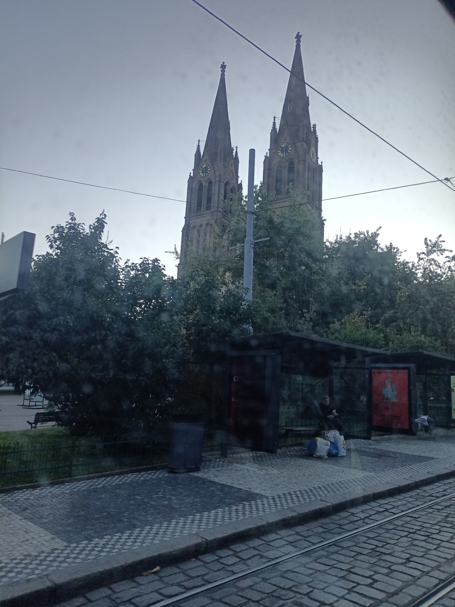 The image depicts an exterior view of the Church of St. Ludmila in Prague, Czechia, with a tram stop in the foreground. Two prominent spires of the church dominate the upper portion of the frame, with visible clocks on their faces. Lush green trees partially obscure the church structure. Below the trees, a modern tram stop shelter is visible, featuring black framing and transparent panels. Inside the shelter, a person is seated, holding a mobile phone. Several shopping bags, appearing to be white and blue, are placed on the ground near the seated person. The ground in front of the tram stop is paved with a checkered pattern of light and dark grey tiles, transitioning to cobblestones in the immediate foreground where tram tracks are laid. A dark cylindrical trash bin is positioned to the left of the tram stop. The sky is overcast, suggesting a cloudy day.