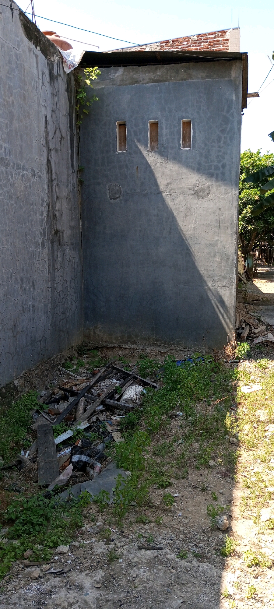 The image shows an outdoor scene in Sengkang, Indonesia, focusing on the corner of a building with a concrete wall. The wall is textured and shows signs of age, with visible cracks. Three small, rectangular windows with wooden frames are present on the wall. A large shadow falls diagonally across the wall, indicating the sun's position. Below the wall, there is a pile of debris consisting of various objects, including wooden planks, broken pieces of material, and possibly some plastic waste, interspersed with green vegetation. The ground is a mixture of dirt and small stones. To the right of the building, a path or open area is visible, with more green vegetation and the trunk of a tree. The sky is clear and bright blue.