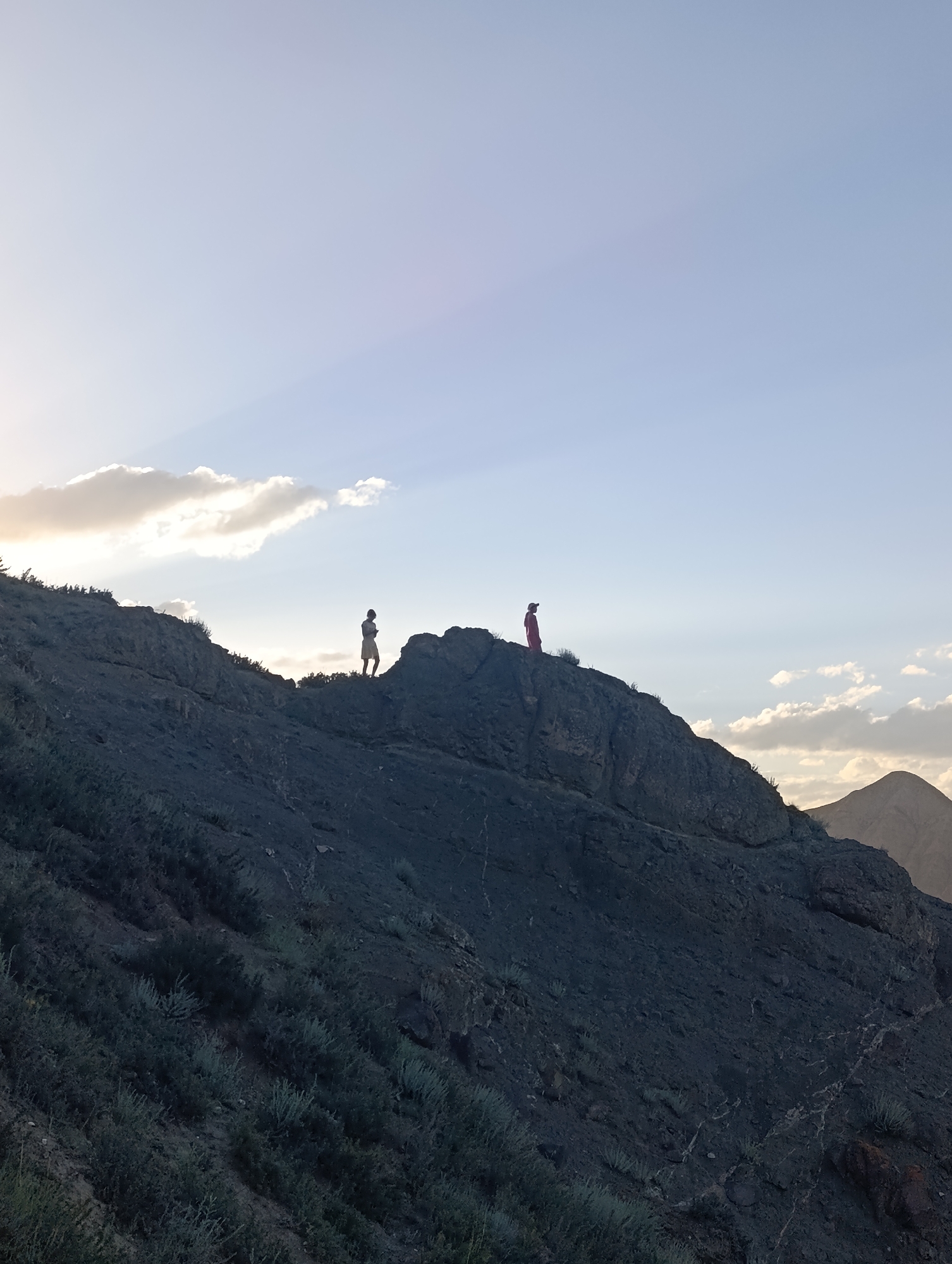 In Naryn City, Kyrgyzstan, two individuals are silhouetted against a pale blue sky on a rocky mountain ridge at dusk. The person on the left, wearing a light-colored garment, stands facing left. To the right, another figure in a red outfit stands facing the viewer. The terrain is rugged and covered with sparse vegetation. The sun is low on the horizon, casting soft light and creating subtle rays through the clouds. In the background, more mountains are visible, softened by the twilight.