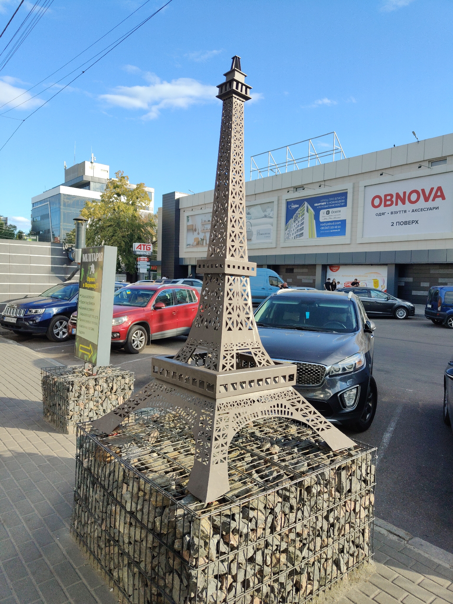 A replica of the Eiffel Tower, made of metal with intricate cut-out patterns, stands on a gabion base filled with stones. In the background, a shopping center complex is visible with multiple stores including "OBNOVA" which sells clothing, footwear, and accessories, and "ATG MARKET". Several cars are parked in front of the shopping center, including a dark SUV, a red SUV, and a blue Jeep. A tall sign with Ukrainian text, indicating "MILITARIA," is positioned to the left of the Eiffel Tower replica. The sky is blue with scattered white clouds. The scene appears to be an outdoor parking area in Cherkasy, Ukraine, likely in front of a commercial establishment.