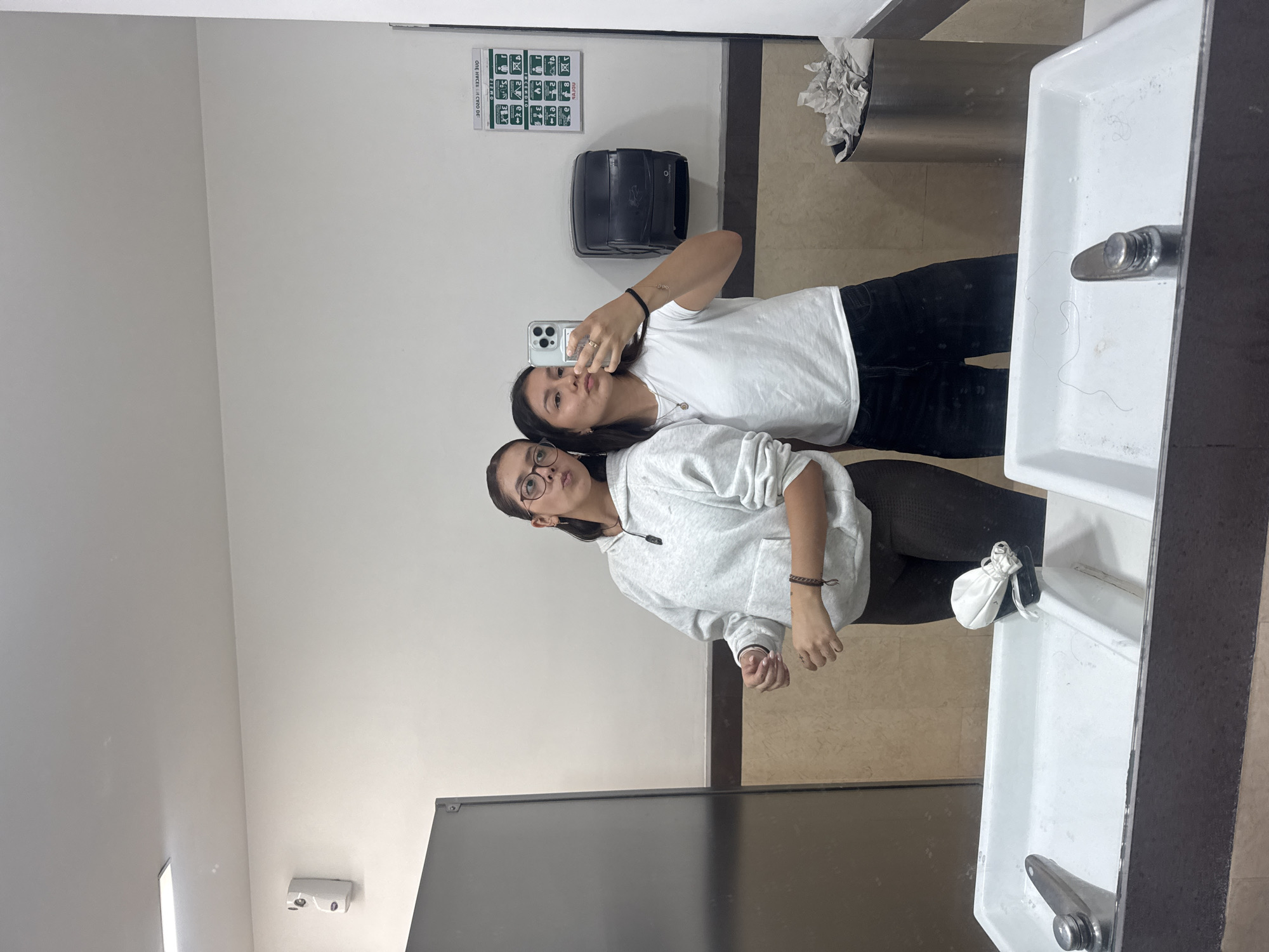 Two young women are captured in a mirror selfie within what appears to be a public restroom or facility in San Andrés Cholula, Mexico. The image, taken by one of the women holding a modern smartphone, shows them posing casually.
The woman taking the photo is taller, with dark hair, and wears a white t-shirt and dark pants. She is making a playful "duck face" directly into the phone camera. Her reflection shows rings on her fingers and a dark bracelet on her wrist.
Standing in front of her is the second woman, who has dark hair, glasses, and is wearing a light grey hooded sweatshirt and dark pants or leggings. She looks towards the camera with a neutral to slightly pouting expression. She also wears a dark, braided bracelet.
The background shows a brightly lit indoor space. Behind the women, to their right (viewer's left), is a white wall featuring a green-and-white instructional sign. Correcting for the mirror's reflection, the text "CICLO DE / ONE WHEEL" is partially visible at the top, followed by rows of green pictograms indicating various safety or instructional procedures (e.g., handwashing, first aid, exit routes). Below the sign is a black paper towel dispenser.
To the women's left (viewer's right), a stainless steel trash can filled with crumpled paper is visible. In the foreground, a white porcelain sink or water fountain with a metallic faucet is prominent, containing a few strands of dark hair. A small white drawstring pouch rests on the edge of the sink. The floor is tiled in a light brown or beige color, with a darker brown counter or partition below the mirror. The overall ambiance suggests a functional, possibly institutional, public space. The women appear to be enjoying a lighthearted moment.