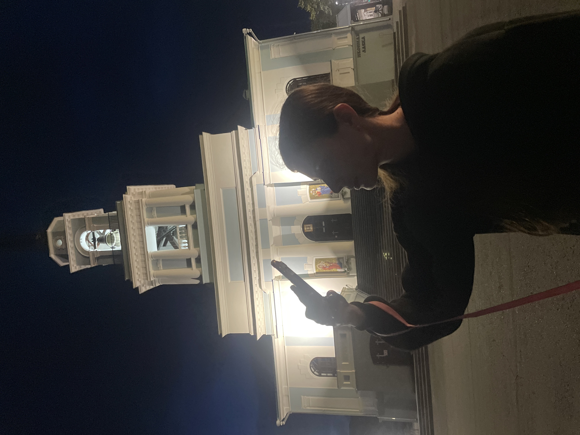A person is captured in profile from the right, with their head bent as they look at their phone. They are wearing a dark top with orange trim on the sleeves. A red ribbon or strap is visible, connected to their wrist and extending out of frame. Behind them, a well-lit white and blue building, possibly a church or administrative building, stands against a dark night sky. The building features a prominent bell tower with an open belfry. The scene appears to be outdoors, with a paved surface visible in the foreground. The lighting suggests it is nighttime. The building has visible text in Cyrillic script above an entrance: "ИКОННАЯ ЛАВКА". This translates to "Icon Shop." The overall mood is one of quiet observation or engagement with technology in a historical or religious setting.