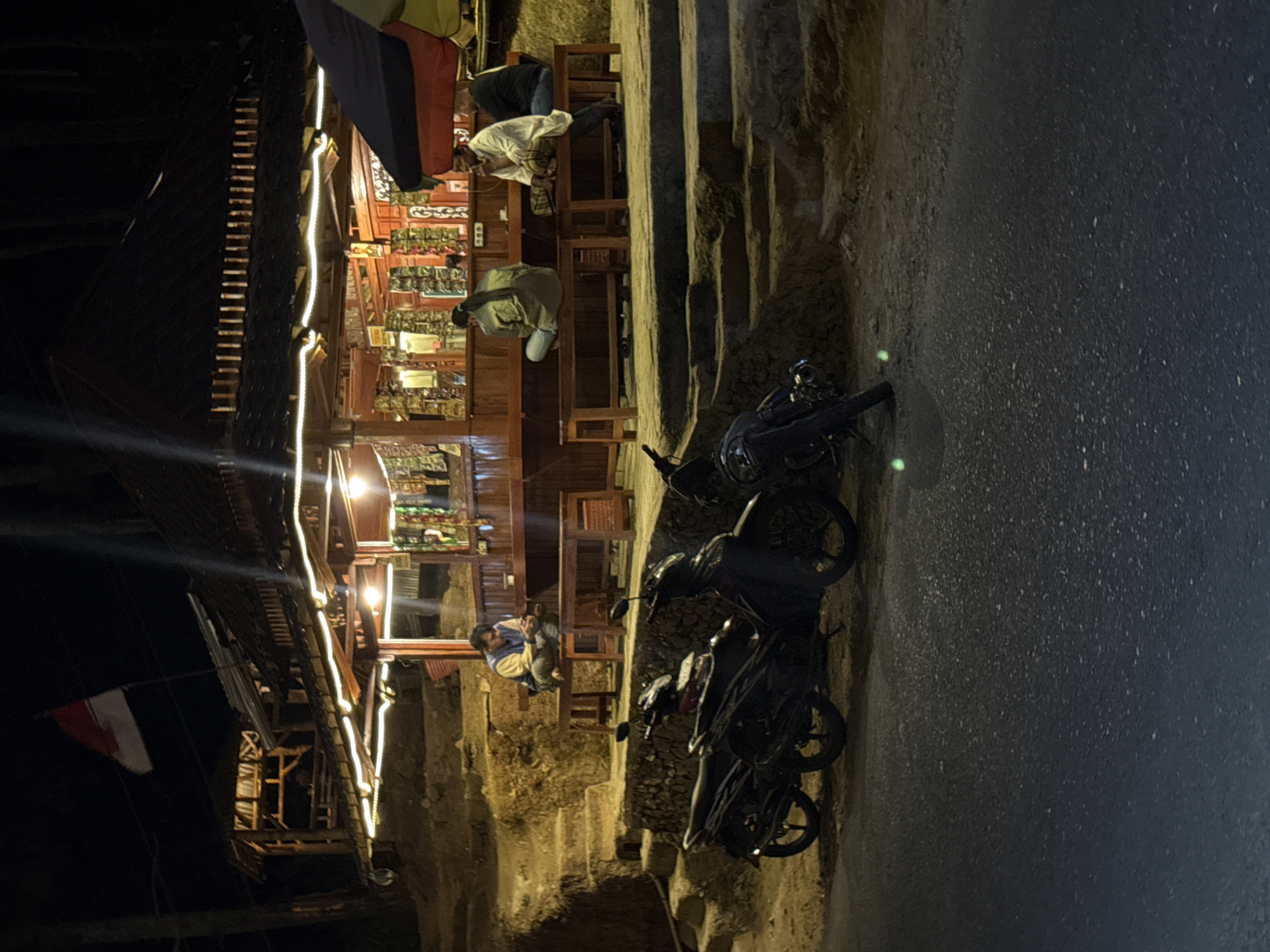 The image captures a nighttime scene at a roadside stall in Slahung, Indonesia. The stall is illuminated by string lights and features a dark wooden roof and counters displaying various packaged goods. Several individuals are seated on wooden benches in front of the stall. A man in a white outfit is seated on the right, facing left. Next to him, a person in a green jacket faces away from the camera. Another individual in a blue and yellow jacket is seated on a bench to the left, looking down at their phone. In the foreground, two motorcycles are parked on the side of the road. The setting appears to be a rural or semi-rural area, with rough terrain and a stone wall visible behind the stall. A red and white flag is visible flying from a pole above the stall.