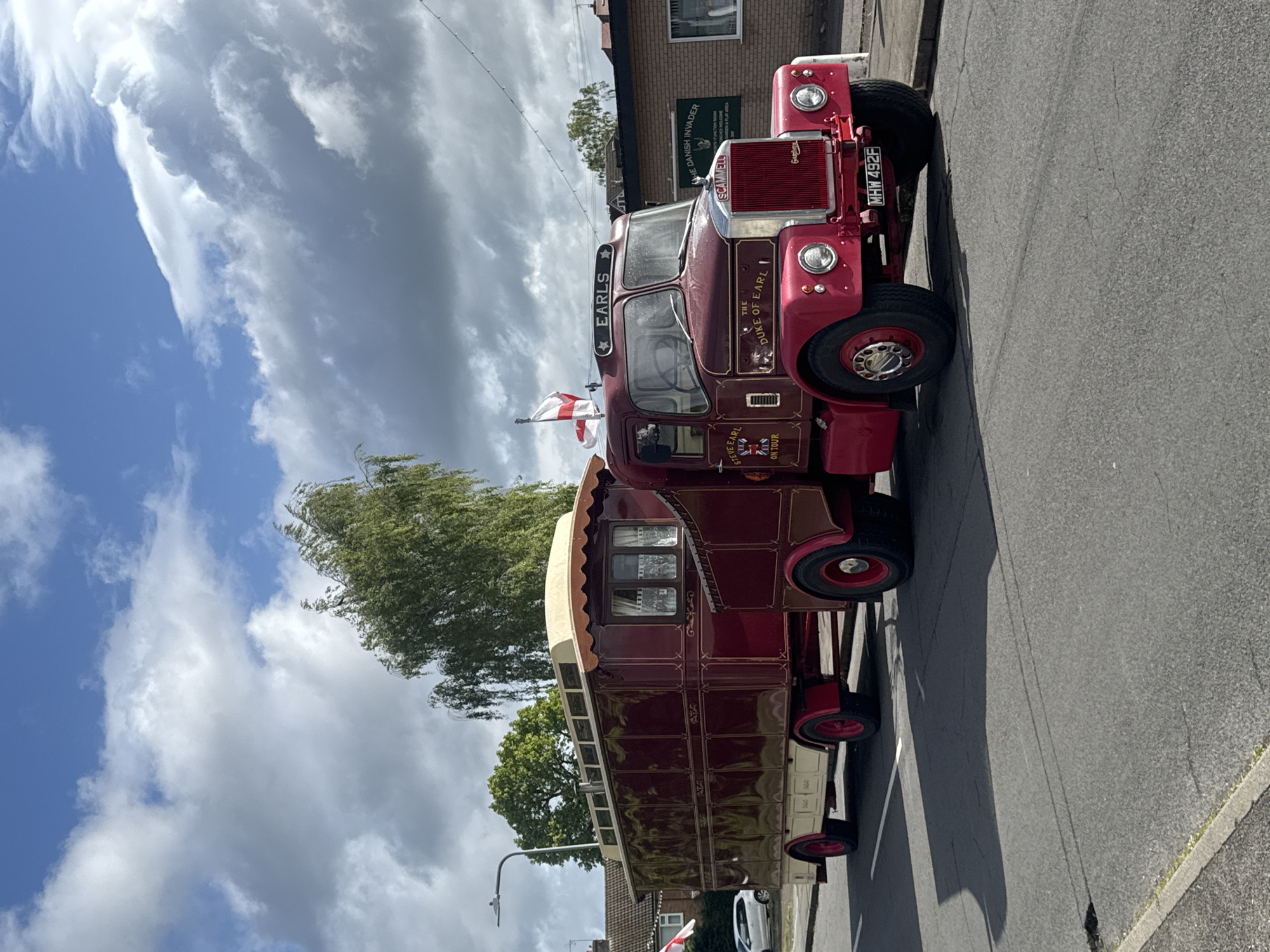 The image captures a maroon and cream Scammell Showtrac, bearing the name "EARLS THE DUKE OF EARL," parked on a street in South Kesteven, United Kingdom. The vehicle, registered as MHW 492F, includes a trailer-like structure attached. The sky is partly cloudy. A brick building is partially visible in the background. A street lamp is visible on the left side of the image. The scene appears static, with no visible people or ongoing actions.