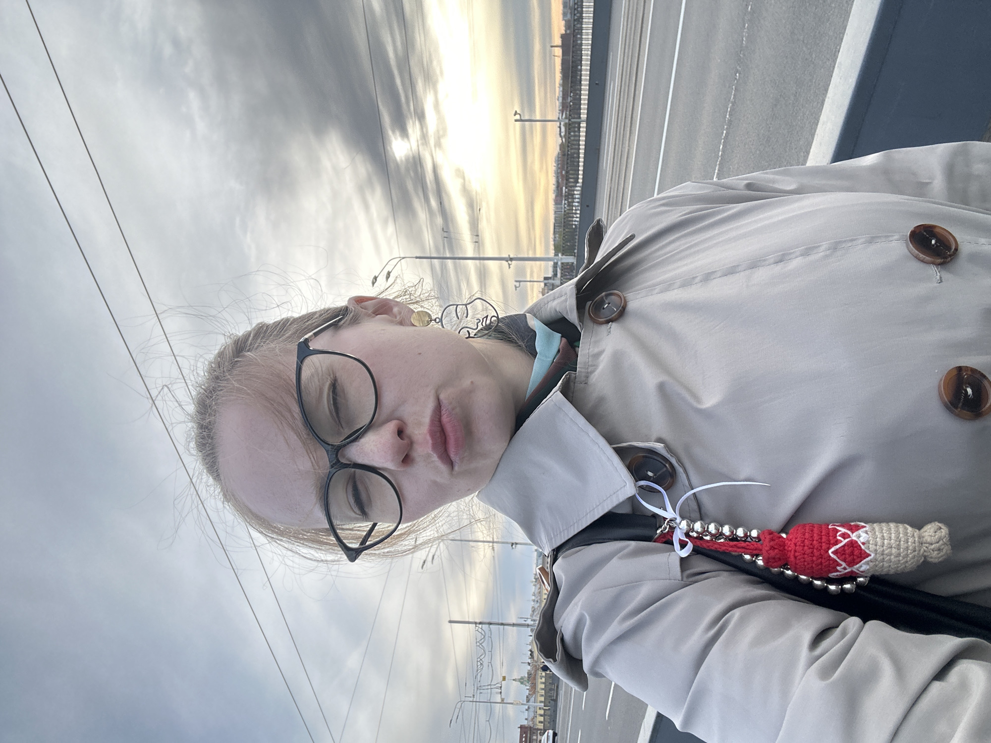 A woman with fair skin, blonde hair, and glasses takes a selfie. She's wearing a beige trench coat with dark buttons. Attached to her coat is a handmade charm, featuring red and beige knitted sections connected by silver beads. The background shows a bridge with streetlights and a cityscape. The location appears to be Saint Petersburg, Russia, given the architecture and bridge design. The sky is overcast with a hint of sunrise or sunset. A highway lane is visible on the bridge.