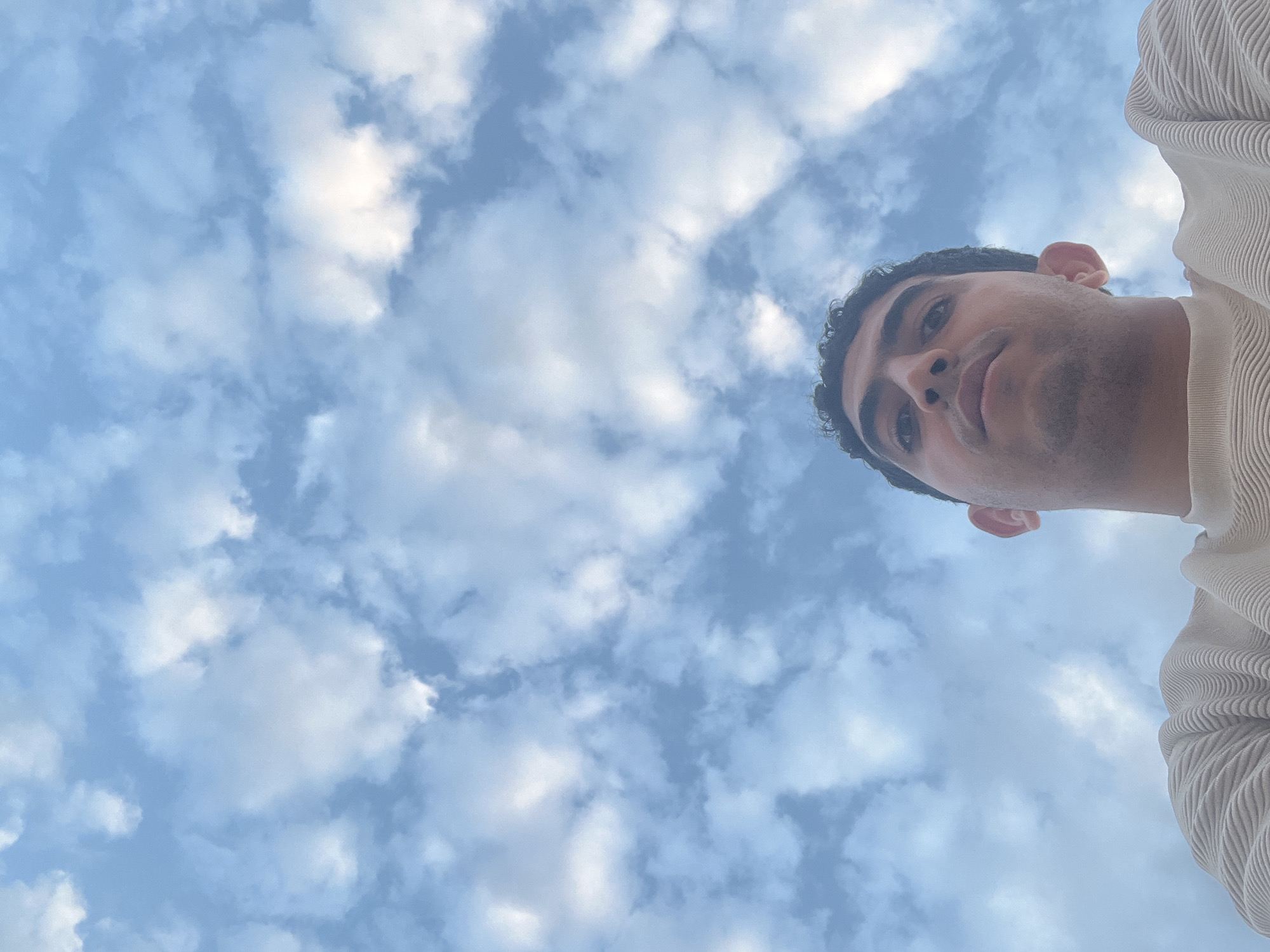 A low-angle, upward-facing photograph showing a person against a cloudy sky. The person, positioned in the upper right portion of the frame, appears to be looking down towards the camera. They have dark hair and are wearing a light-colored textured shirt or sweater. The background consists of a blue sky filled with puffy white clouds. The image was not taken in the city of Ouaouizert, Morocco. The location of this photograph cannot be determined from the image alone.