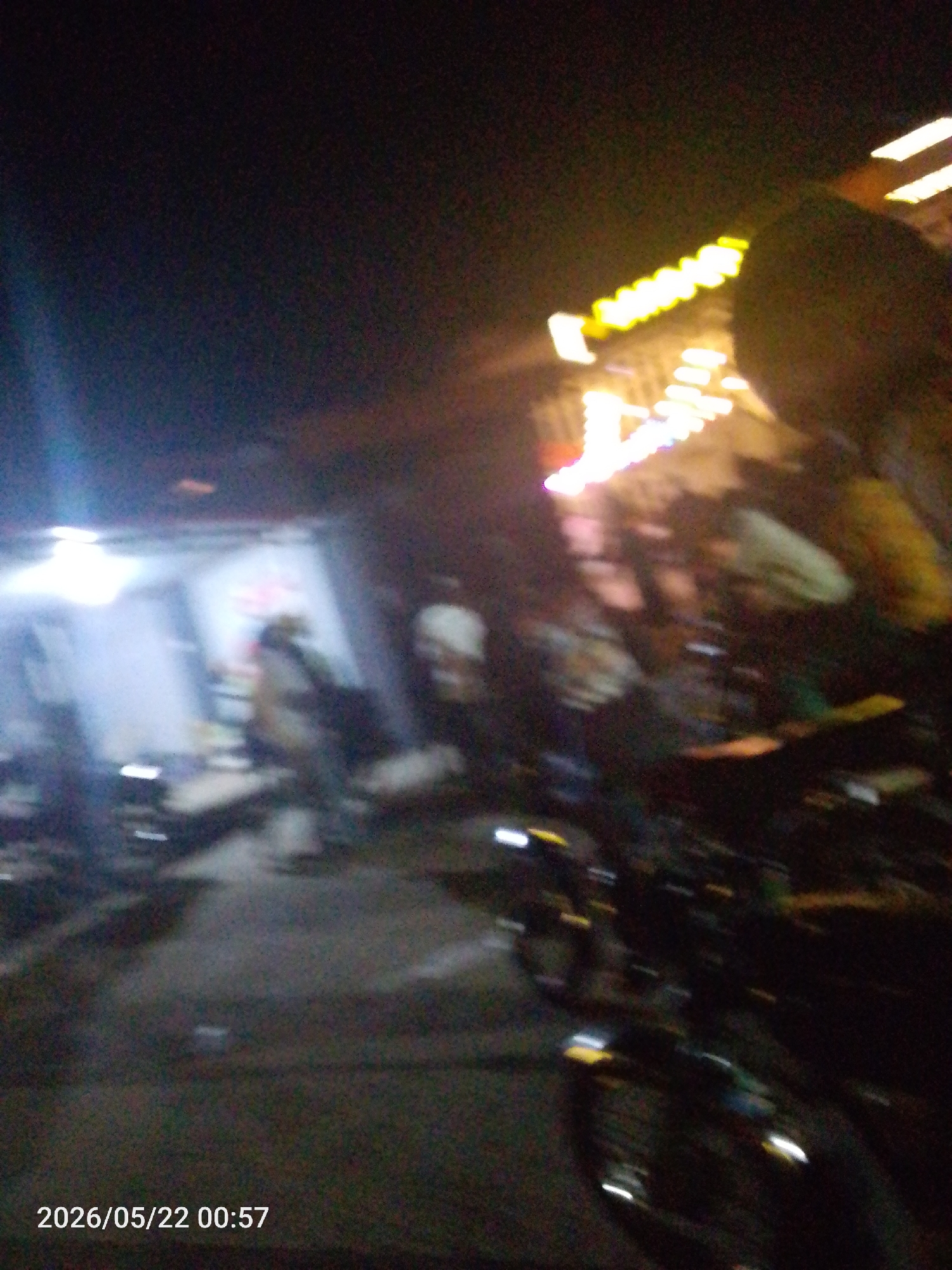At night in Lokoja, Nigeria, the photo captures a blurry street scene. A motorcycle is visible in the foreground. A building with a bright light is visible in the background. A lit sign is above the building. There are blurred figures of people near the building.