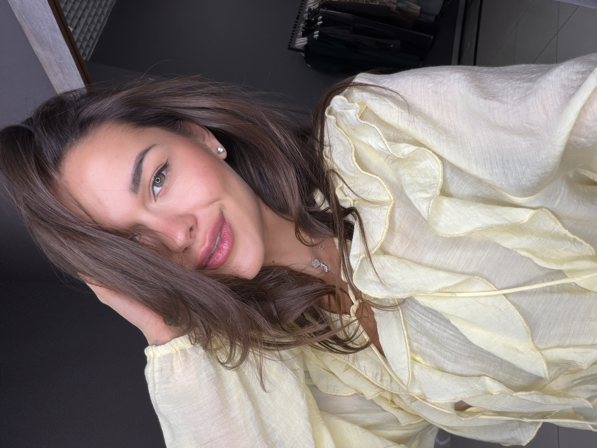 A woman is pictured from the upper chest up, smiling and looking at the viewer. She has light brown hair, visible eyes, full lips, and wears a single earring. She is wearing a light yellow, ruffled, button-down shirt with a necklace visible at her neck. The location is unknown, but based on the prompt, it may be in Krasnoyarsk, Russia. There are clothes hanging behind her on a rack.